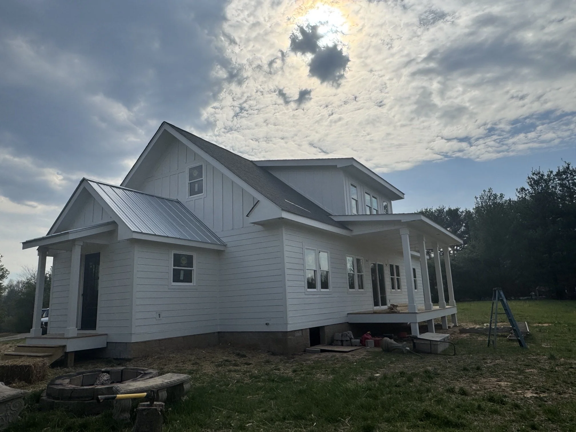 A white two-story house under construction with a partly cloudy sky above, surrounded by some trees and construction materials on the ground.