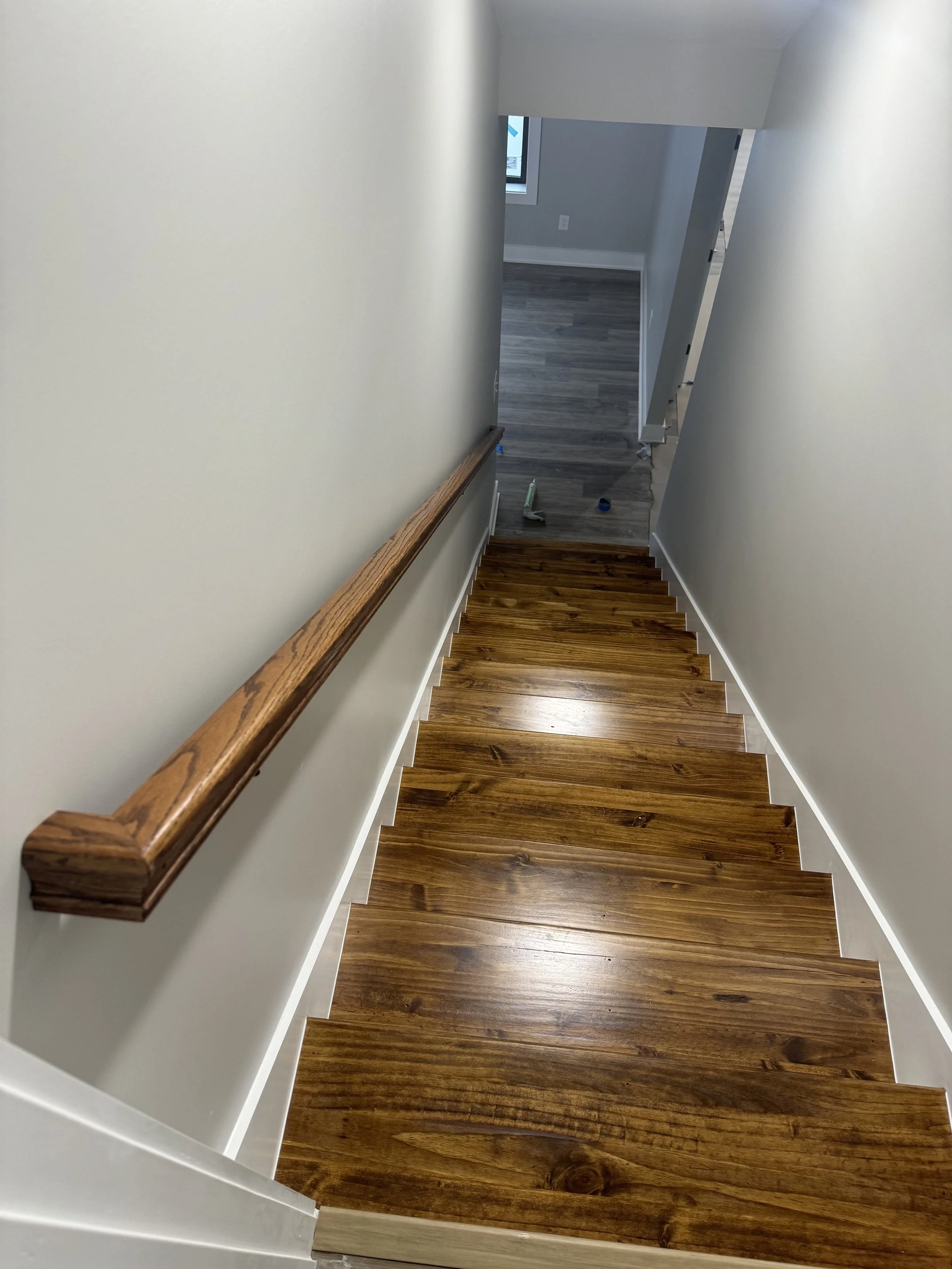 View of a staircase with wooden steps and a wooden handrail, leading down to a room with gray flooring and walls, a window, and some painting tools on the floor.