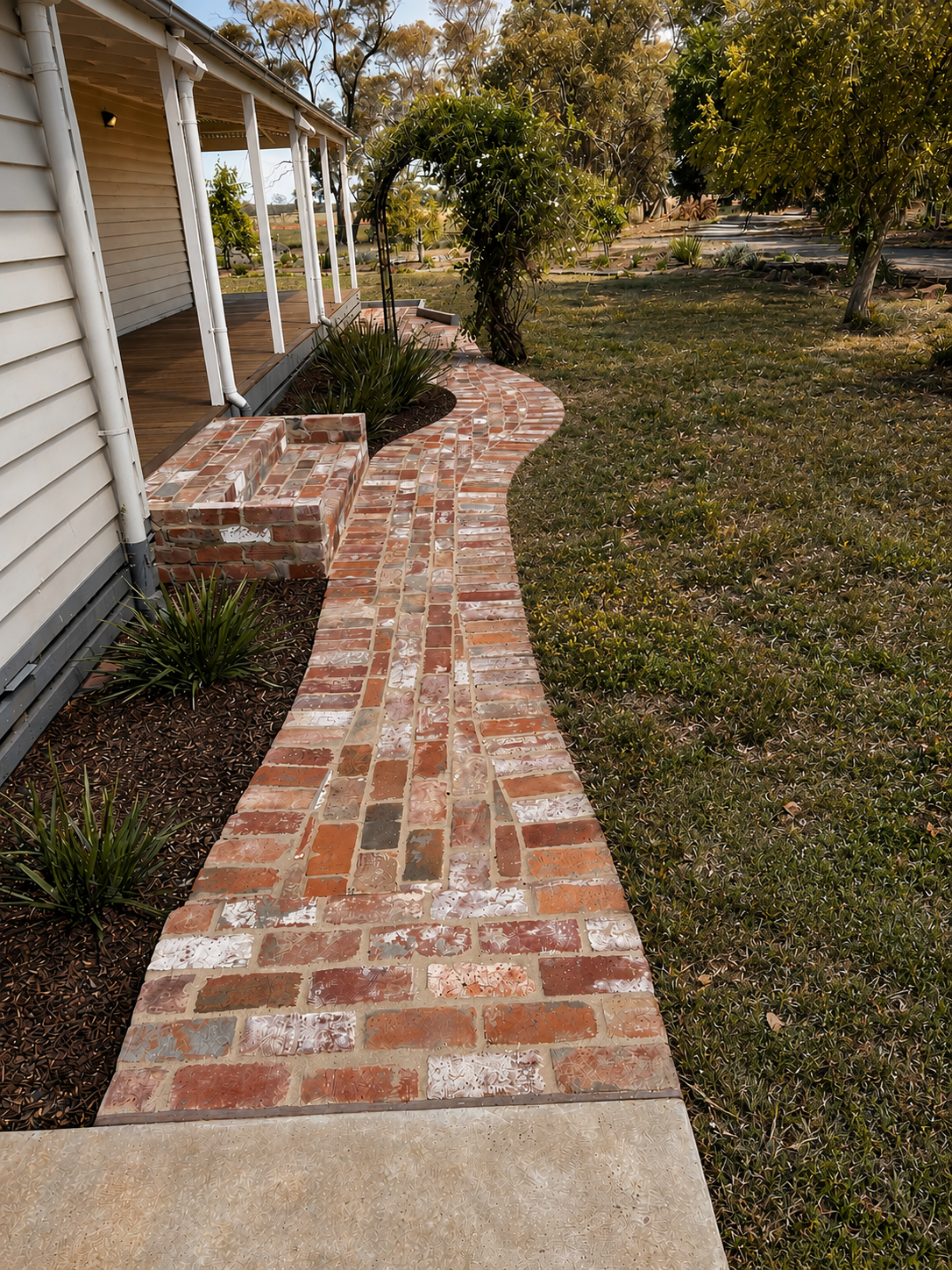 Curved reclaimed brick pathway through established garden beds leading to a weatherboard home.