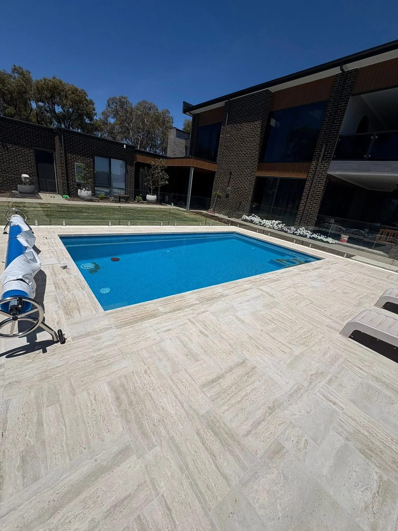 Swimming pool surrounded by travertine stone French pattern paving and landscaped garden beds.