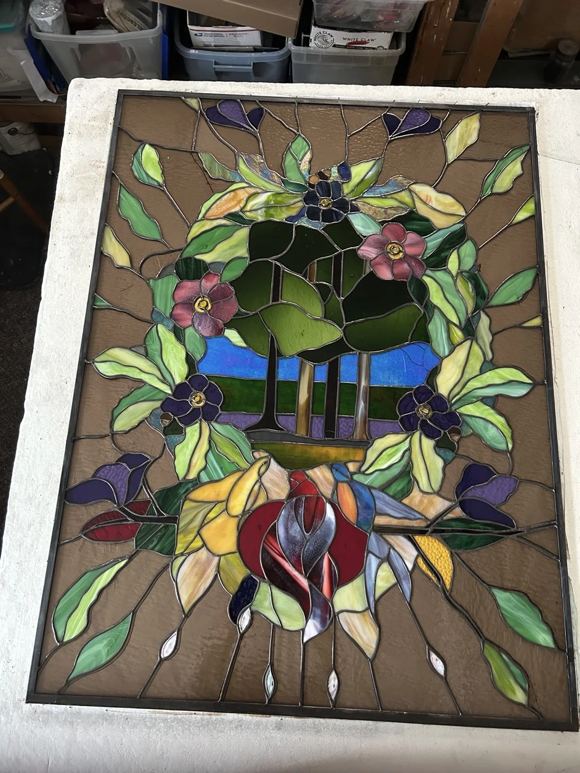 A stained-glass window in progress on a worktable, with green leaves, pink and purple flowers, and a central landscape motif.