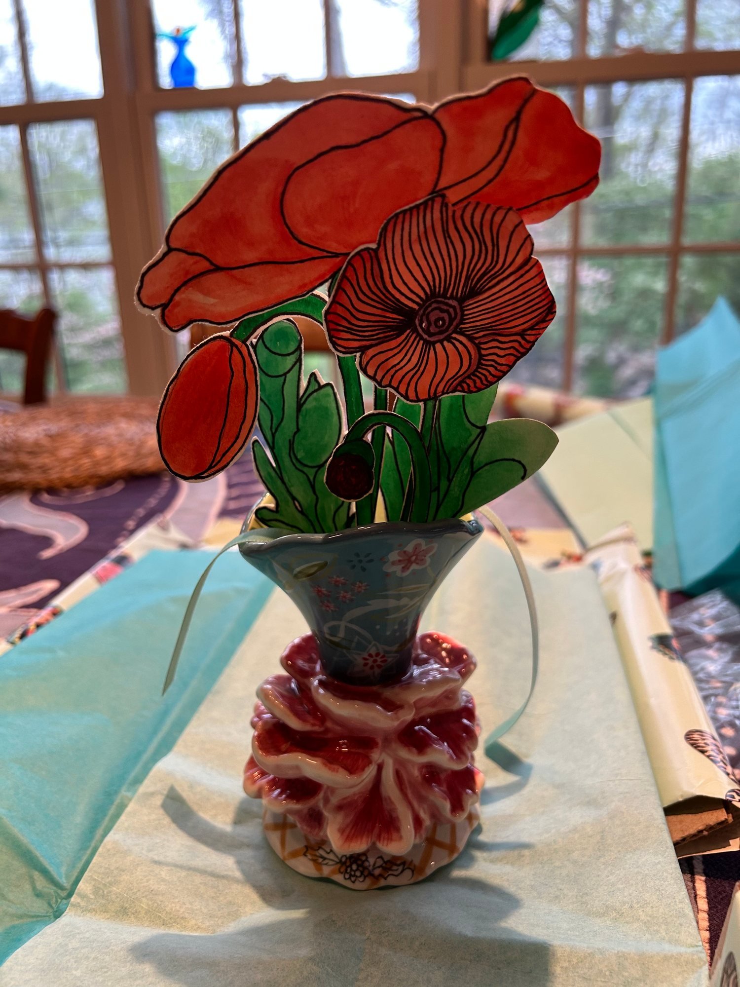 Hand-colored cut-paper poppies in red and orange arranged in a small decorative vase.