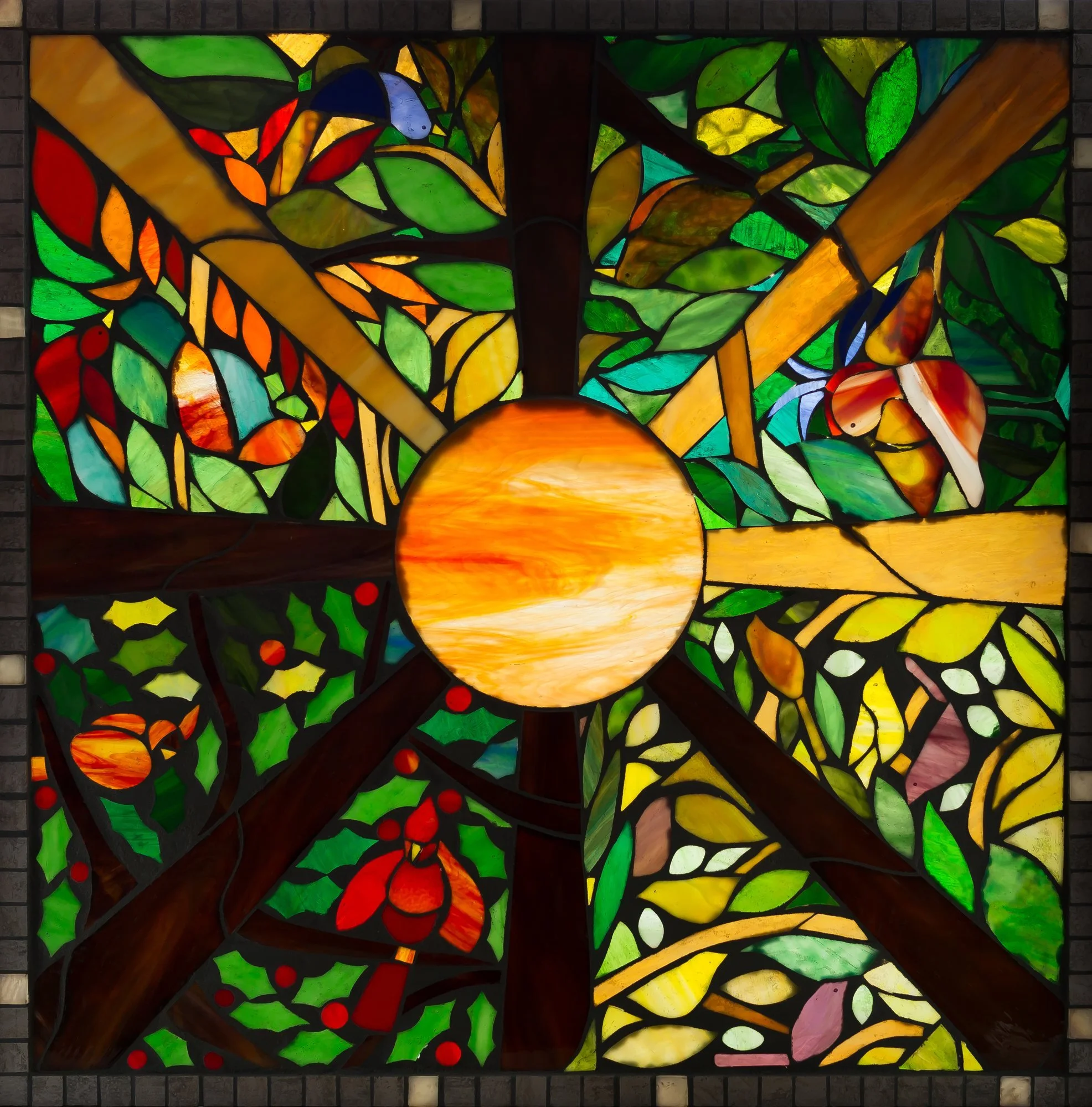 A square stained-glass design with branches and green leaves surrounding a glowing orange circle.