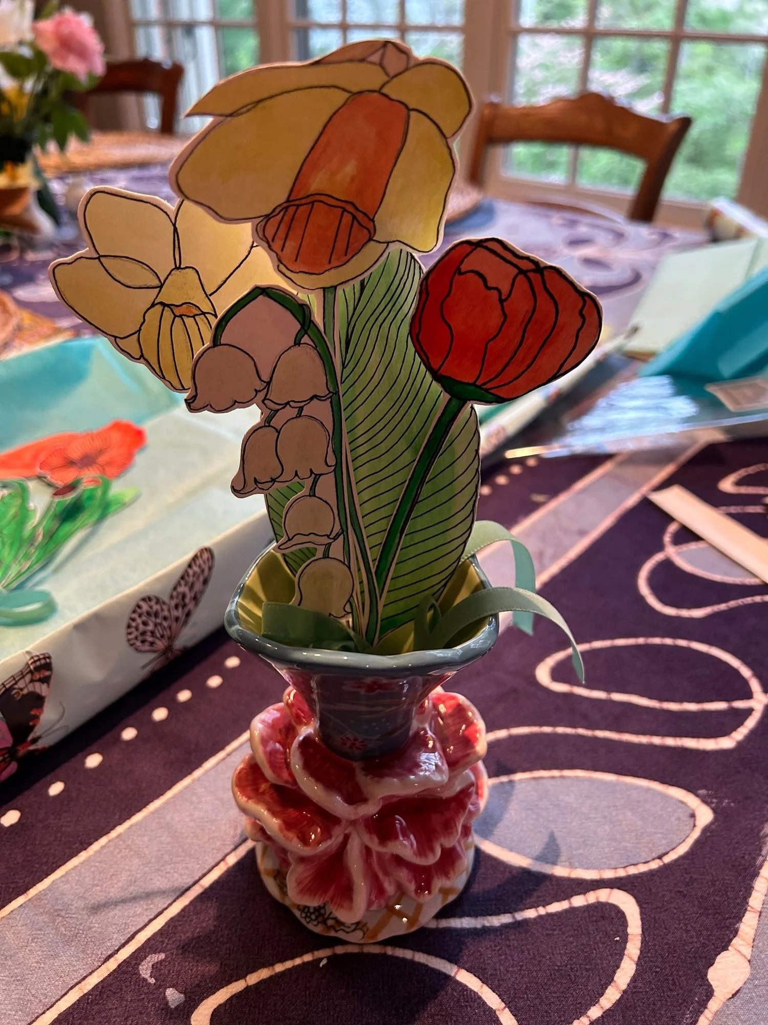 Hand-colored cut-paper daffodils, a red flower, and pale bell-shaped blossoms arranged in a decorative vase.