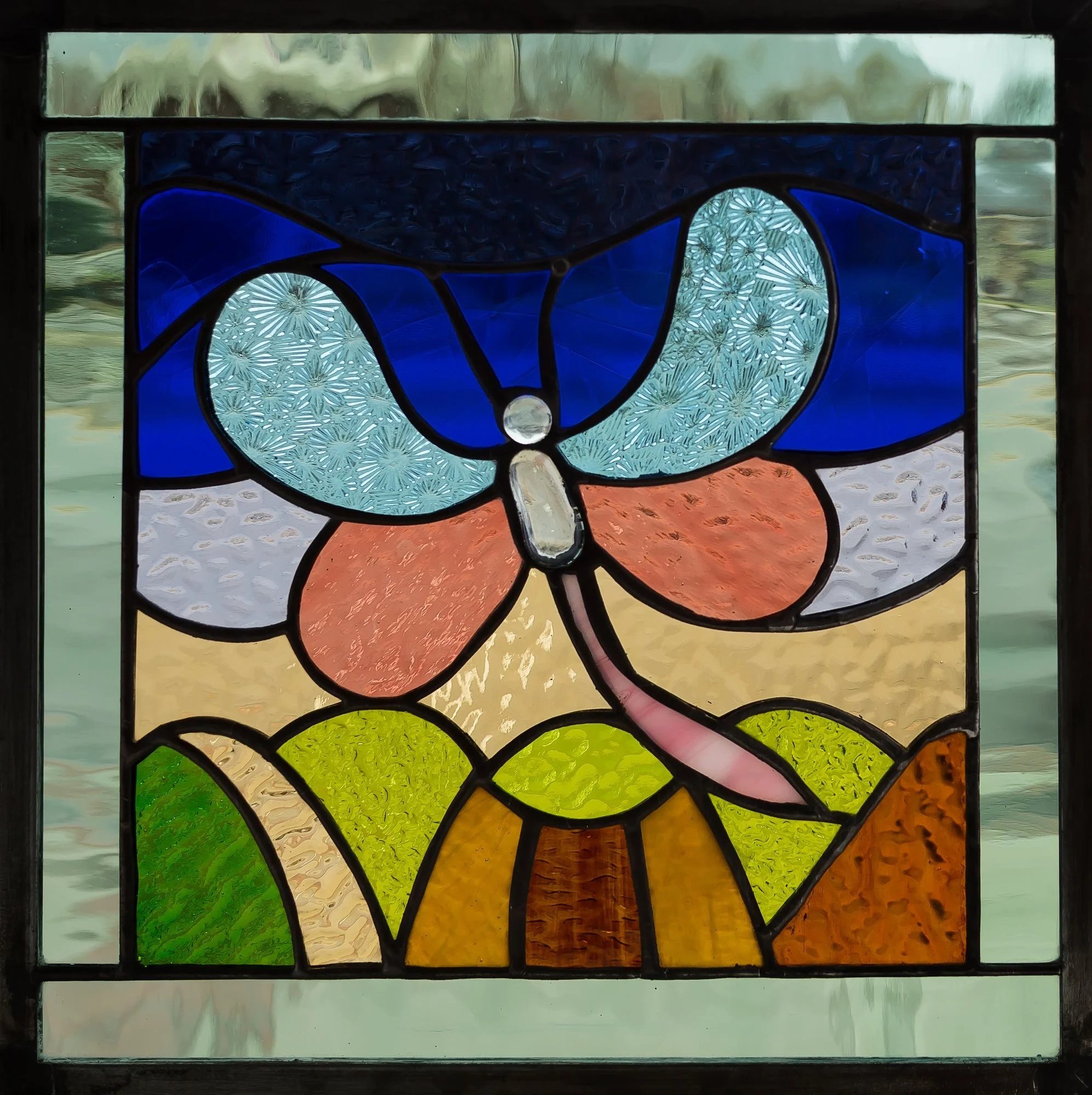 A framed stained-glass butterfly with blue and peach wings over green leaves.