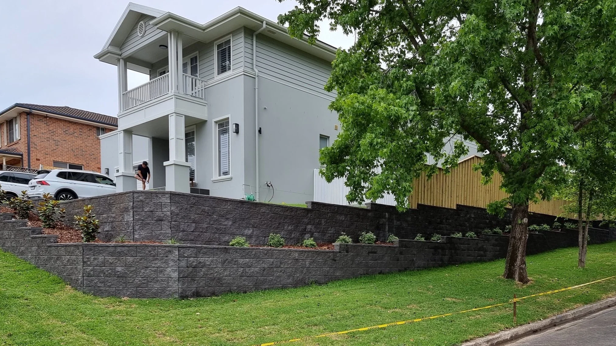 A modern two-story white house with a small porch, surrounded by a landscaped yard with a retaining wall and green trees.