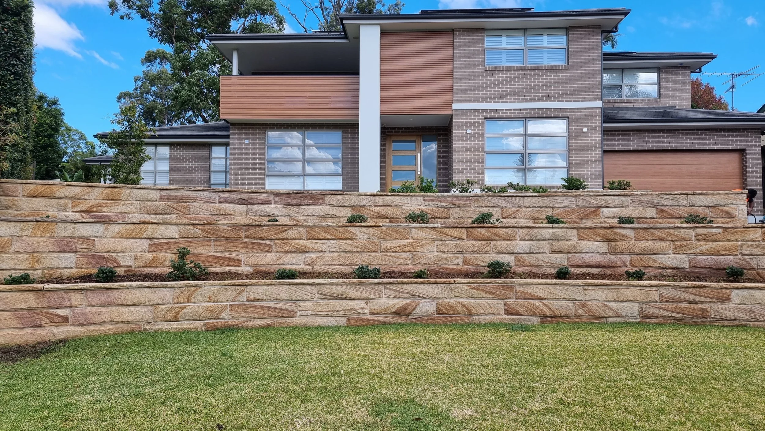 Modern Brisbane multi-story house with brick and wood siding, large windows, and a stone wall with small plants in front, under a blue sky with clouds.