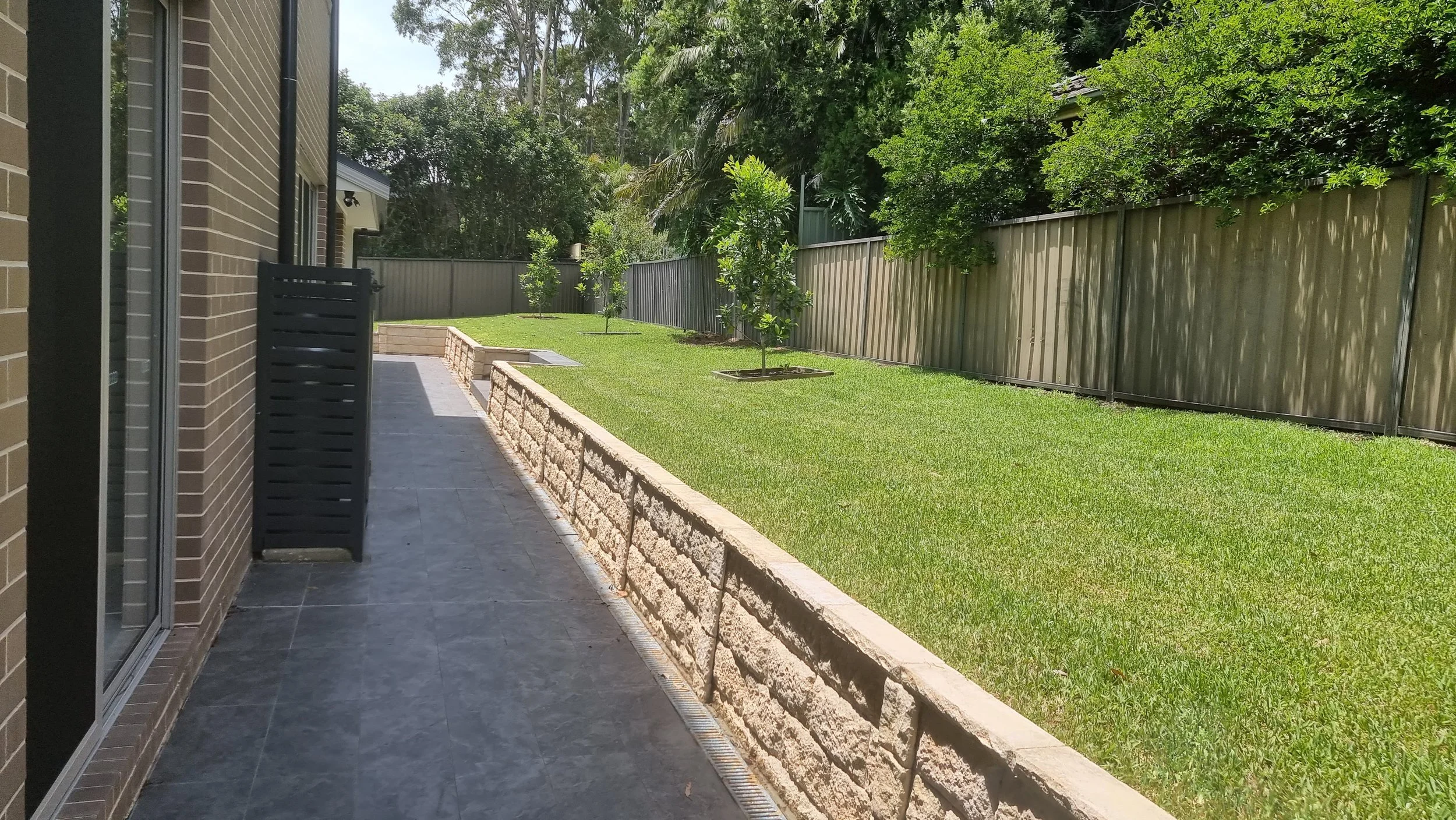 Side yard with grassy lawn, small trees, brick planter wall, metal fence, and paved patio area beside a brick house.