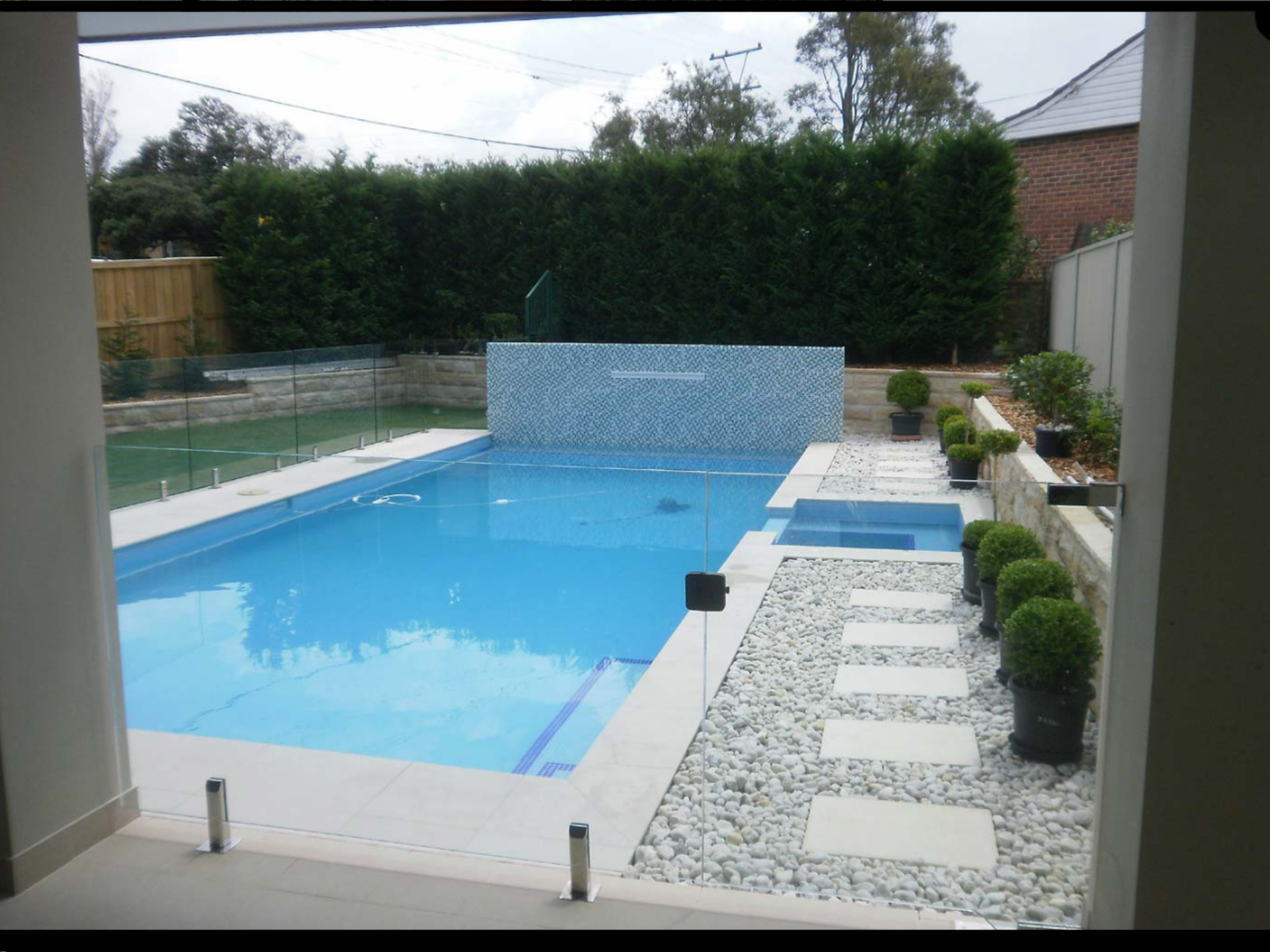 View of a backyard swimming pool with a glass fence, potted plants, and a pebble pathway, surrounded by a brick wall and trees.