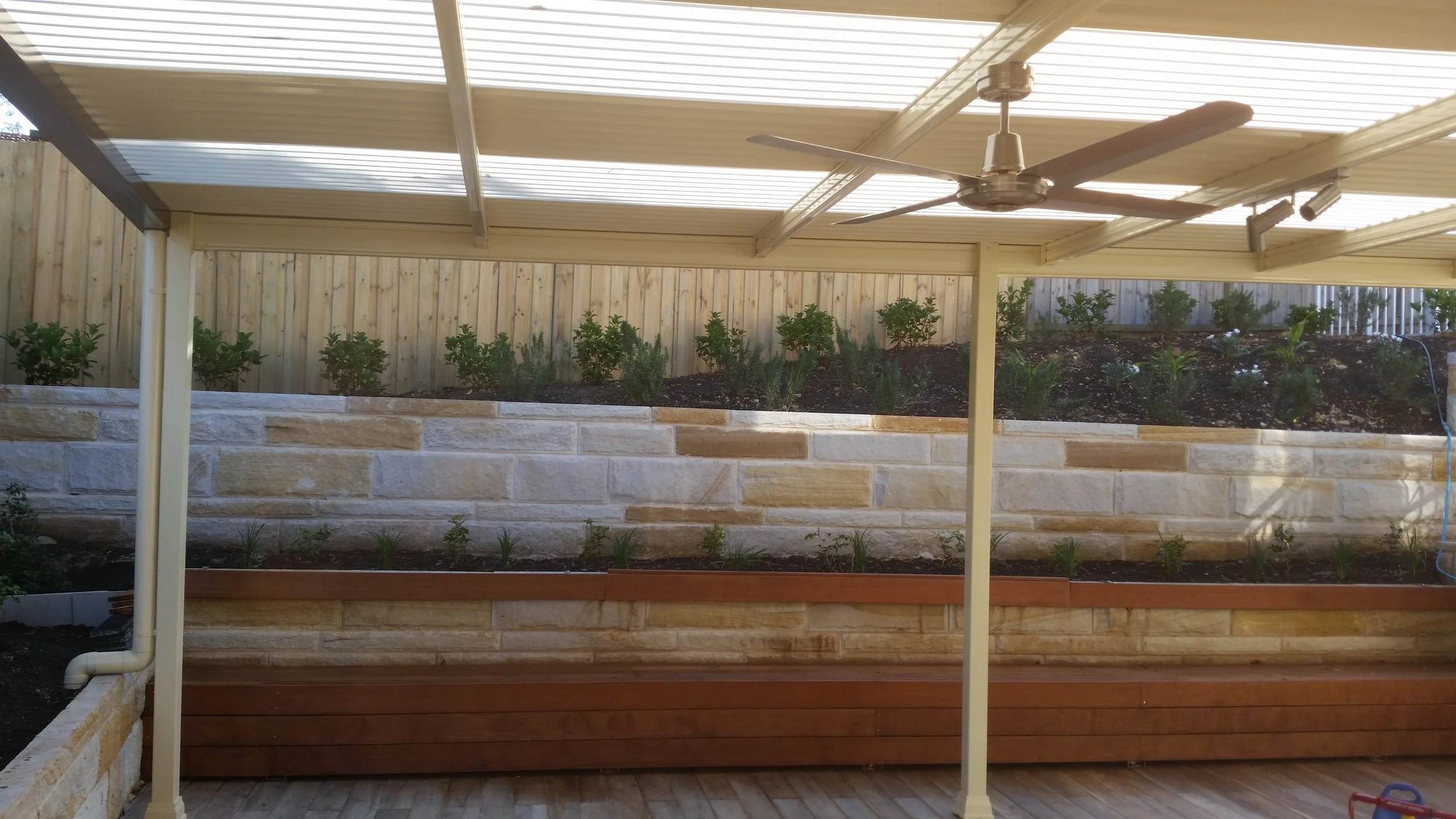 Covered outdoor patio with ceiling fan, wooden flooring, a raised garden bed with small plants, and a wooden privacy fence in the background.