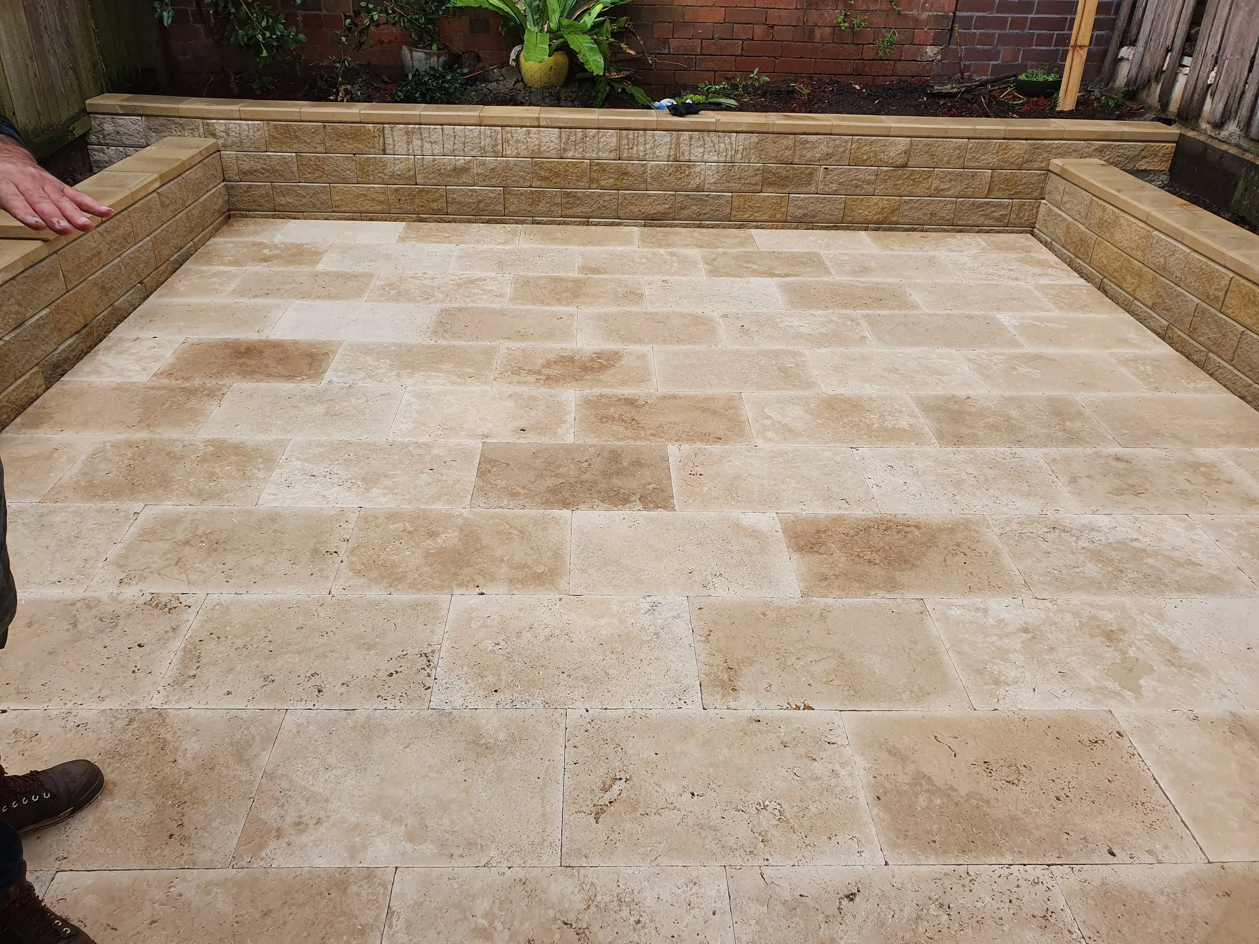 Newly installed beige stone patio with retaining walls and small garden beds in a backyard.