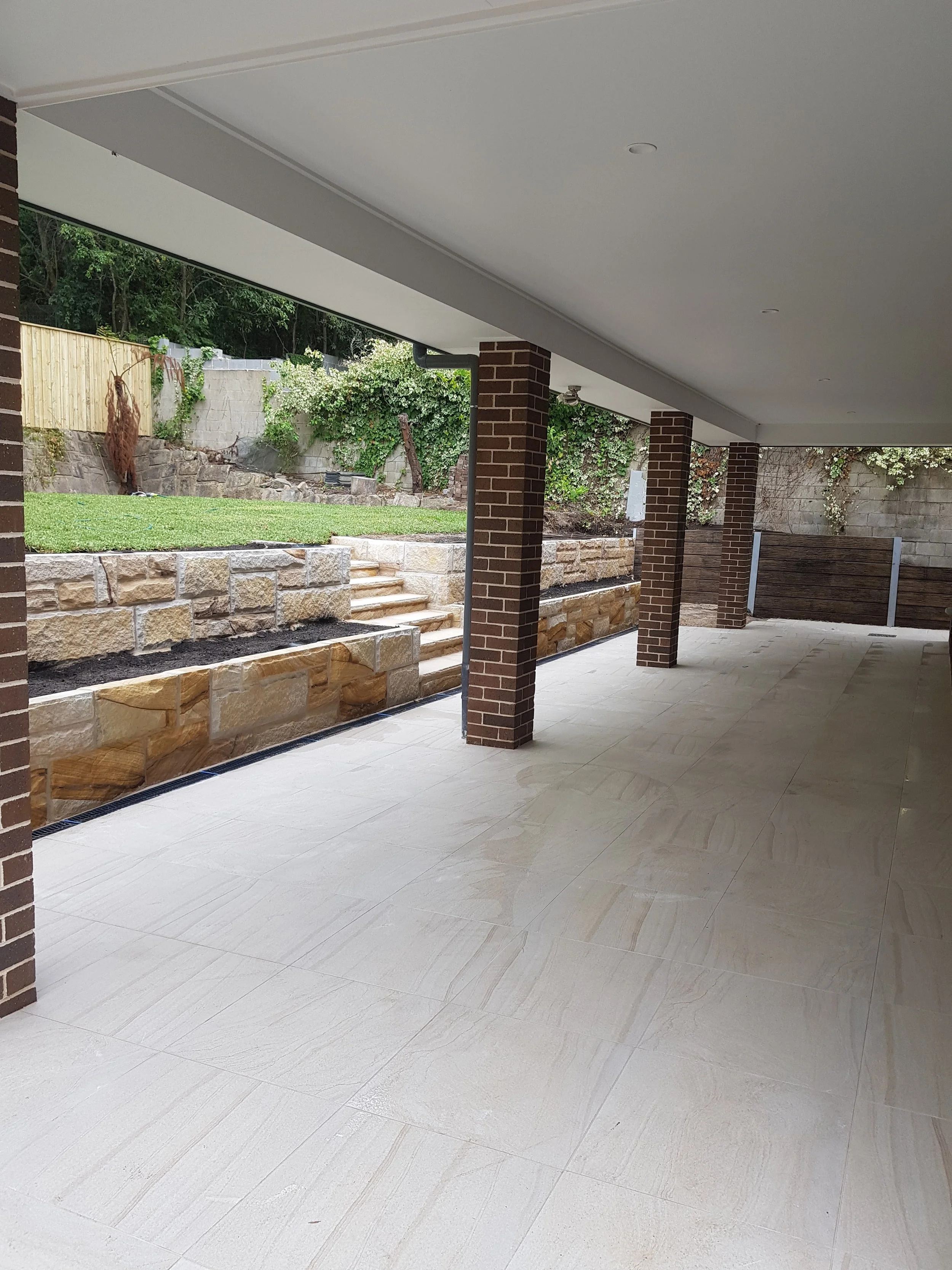 Covered outdoor patio with brick columns, tiled floor, and a view of a tiered garden with stone walls and steps, grass, and plants.