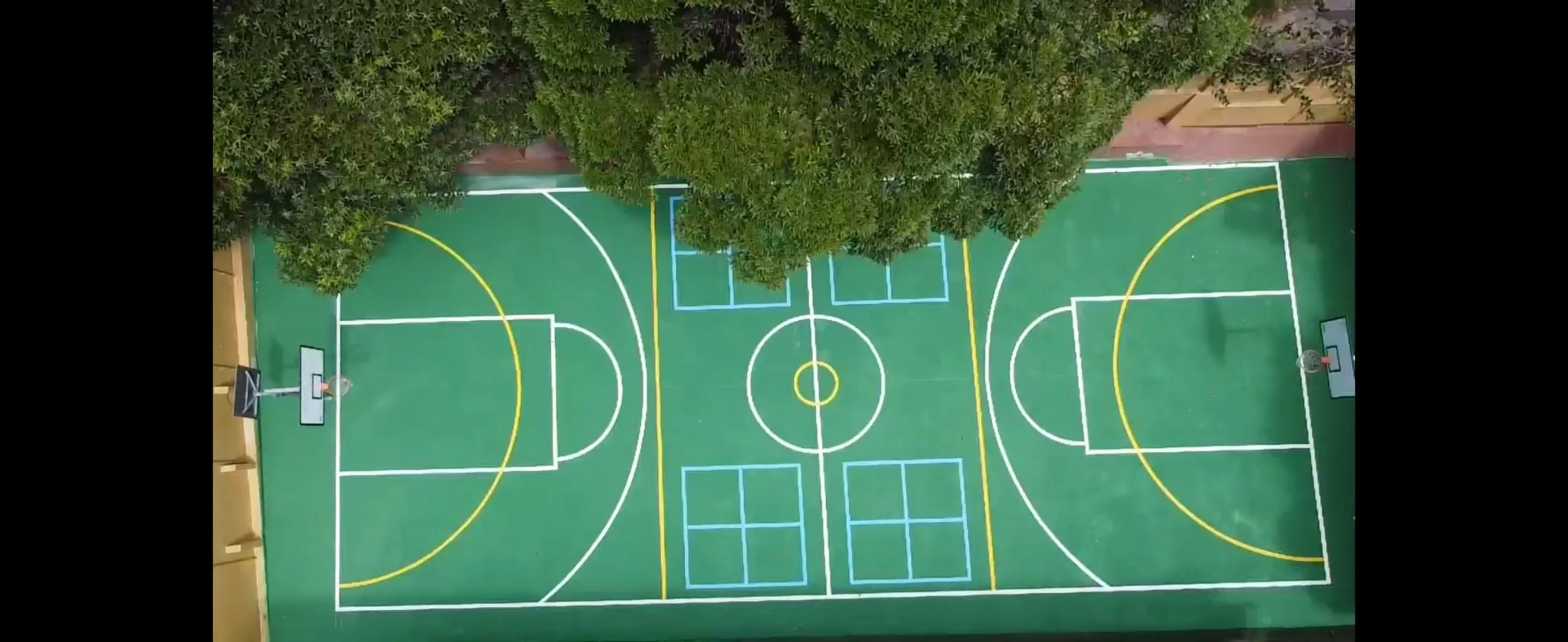 Top-down view of a colorful outdoor basketball court with white, yellow, and blue lines, surrounded by greenery and tall bushes.