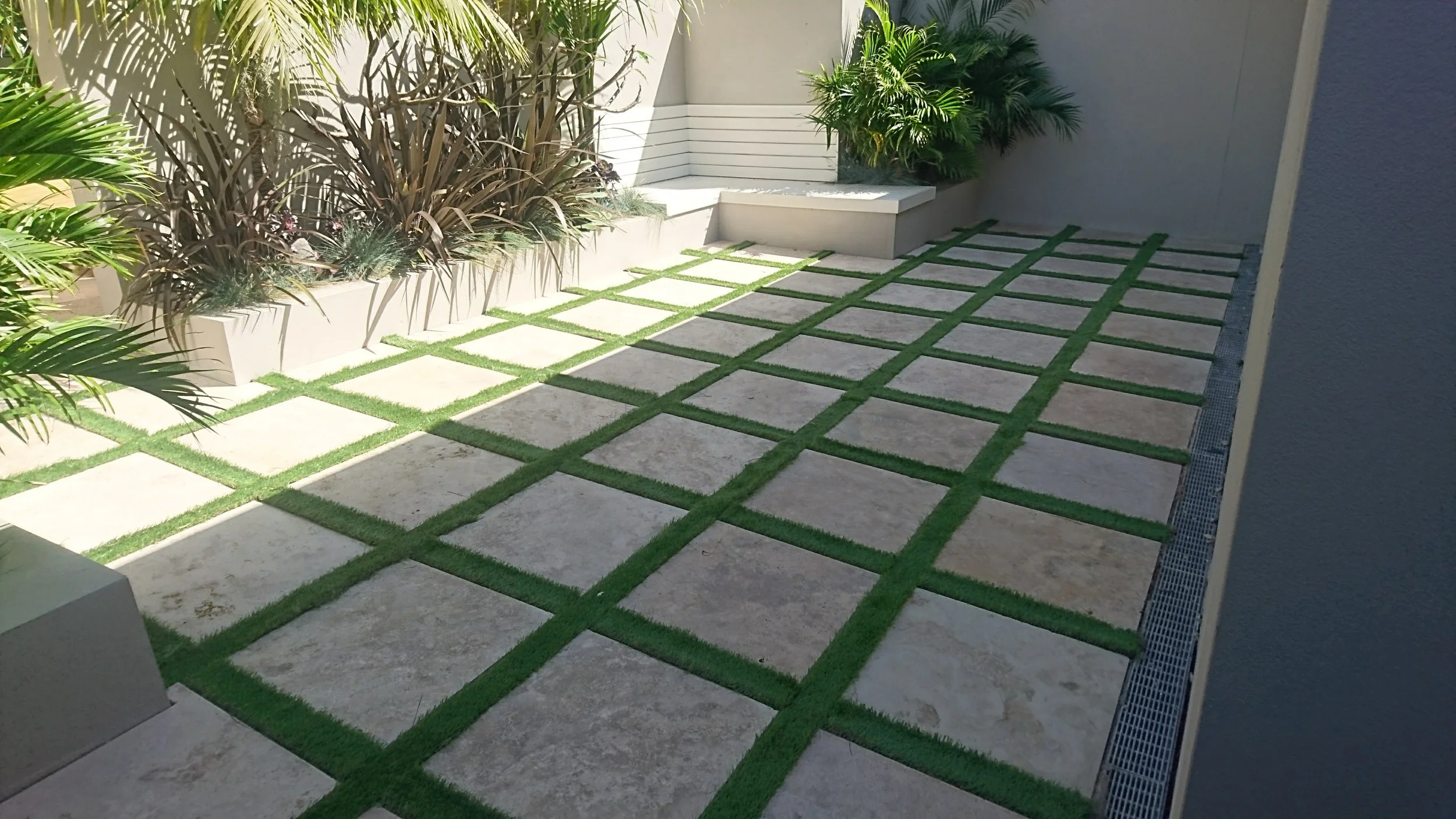 Modern outdoor patio with square stone tiles arranged in a grid pattern, separated by strips of green artificial grass. There are white planters with tall leafy plants along the wall and a small corner bench with potted plants nearby.