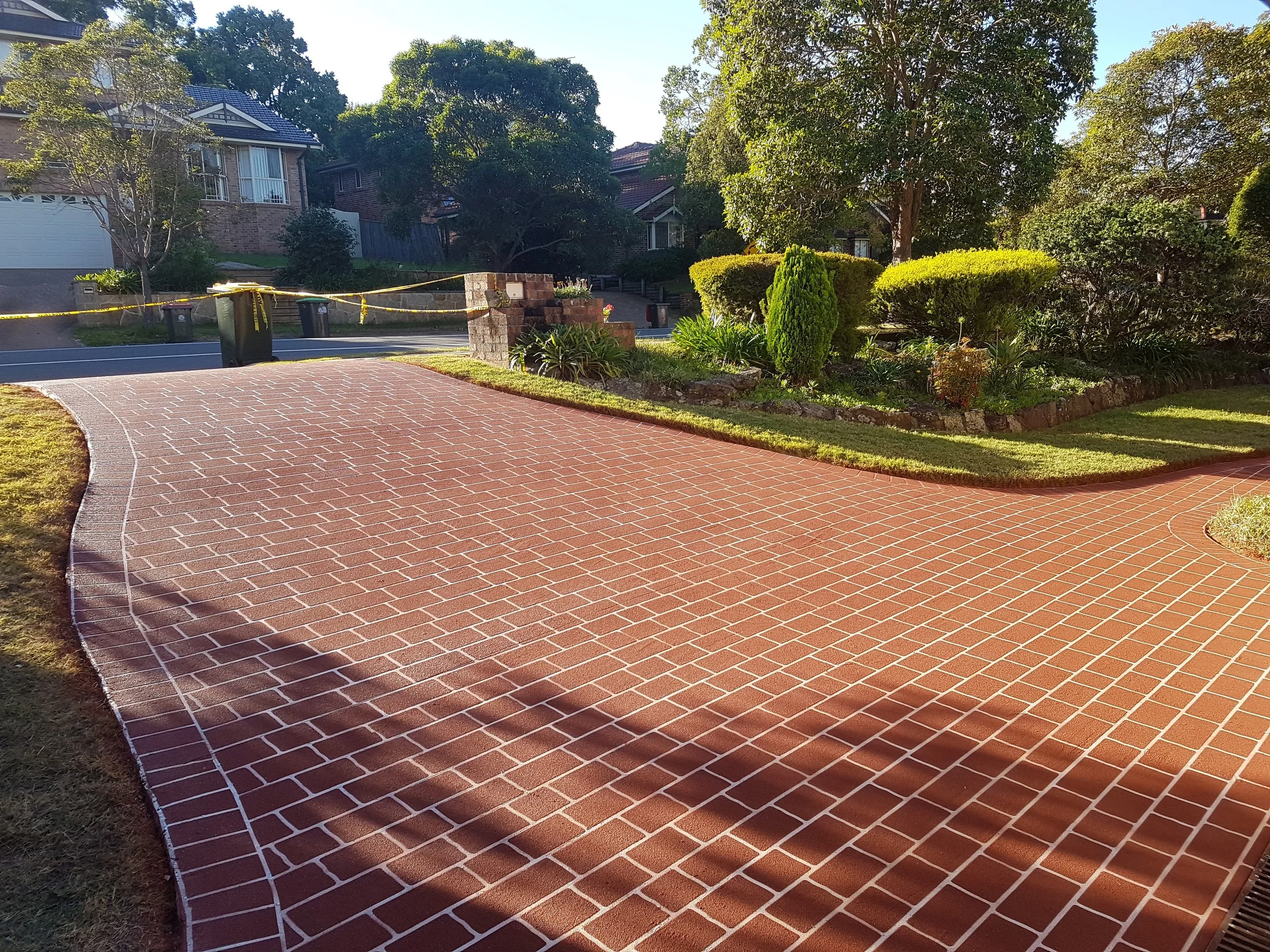 Clean red brick pathway winding through a well-maintained garden area with trimmed bushes and trees, in a suburban neighborhood under clear skies.