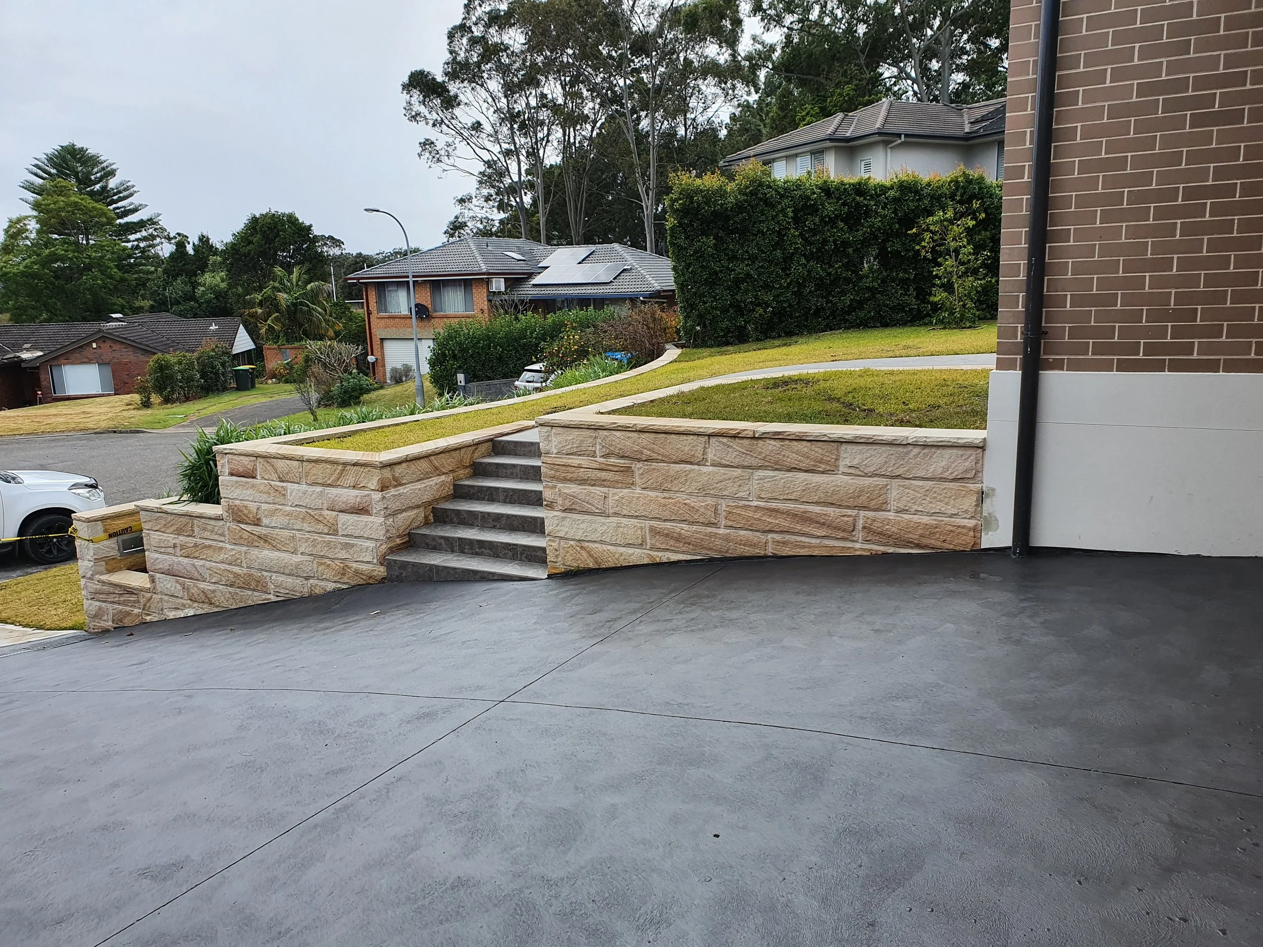 View from inside a garage showing a stone staircase leading to a landscaped lawn and street in a suburban neighborhood with houses, trees, and a cloudy sky.