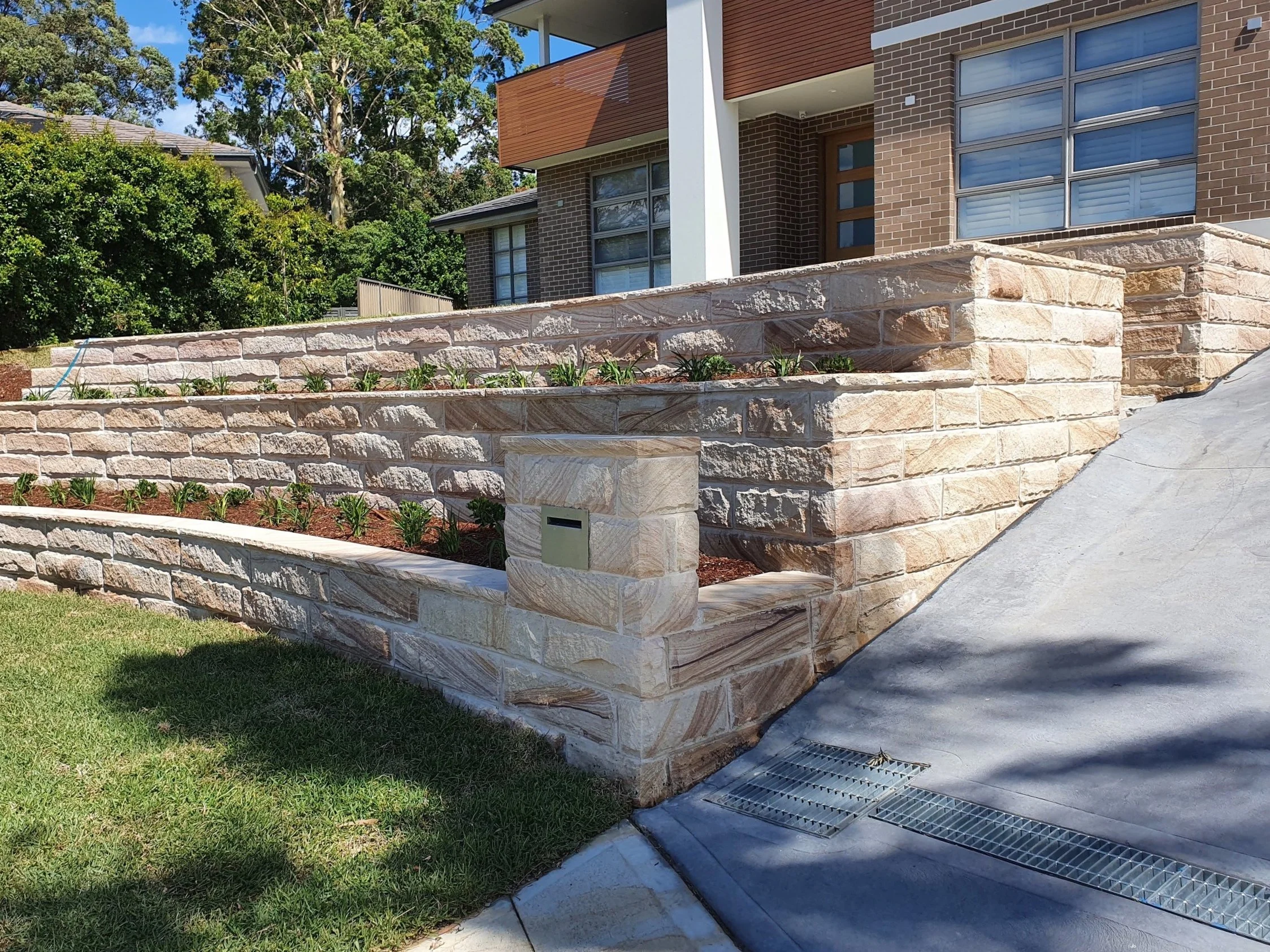 View of a modern house with a landscaped front yard featuring a tiered stone retaining wall, new plants, and a mailbox. There is a concrete driveway with drainage grates and green grass.