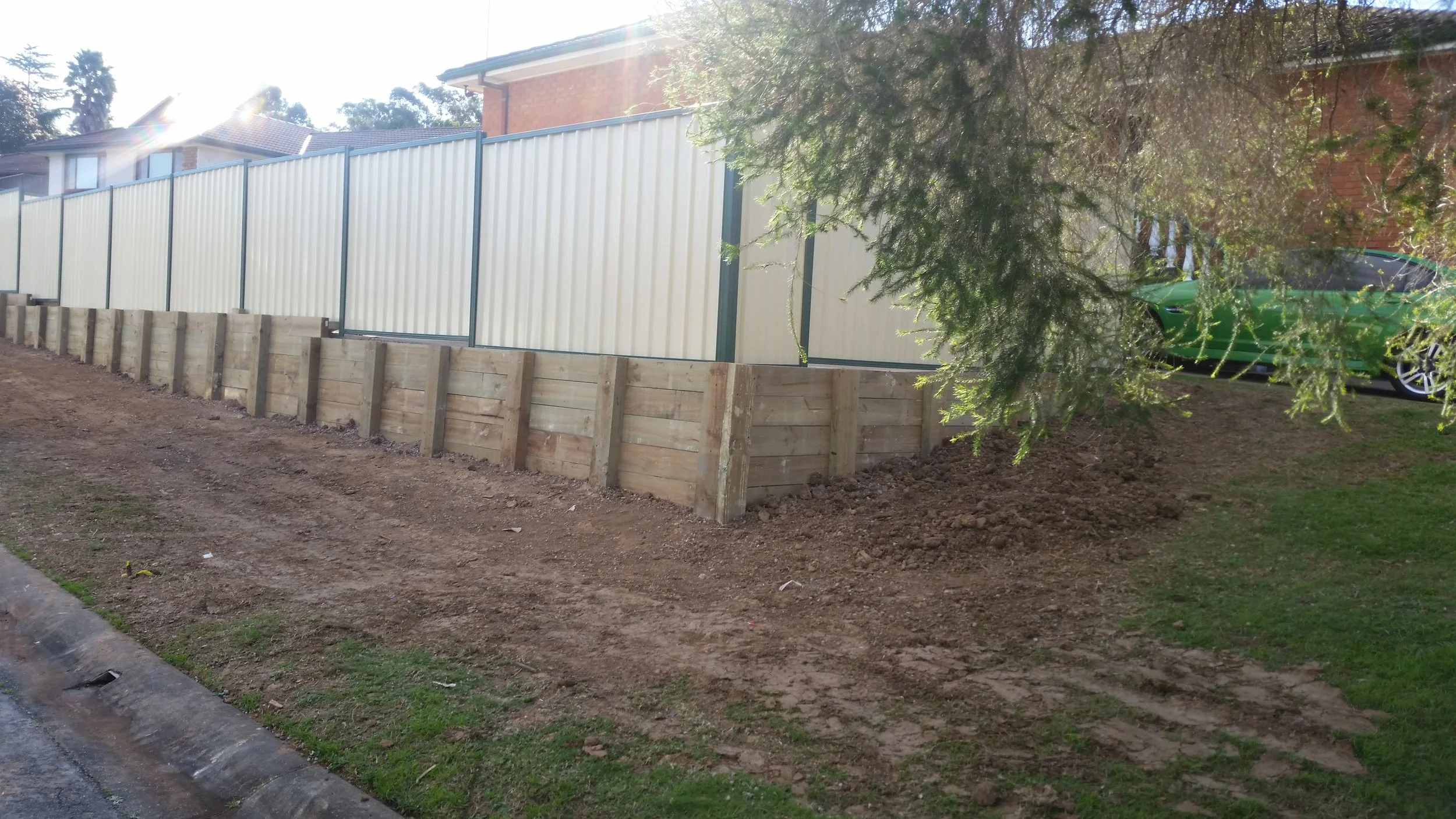 A construction site with a new wooden retaining wall adjacent to a beige metal fence. There are tire tracks and dirt on the ground, a green car is parked behind the fence, and a large tree partially obscures the view, with sunlight shining from the l