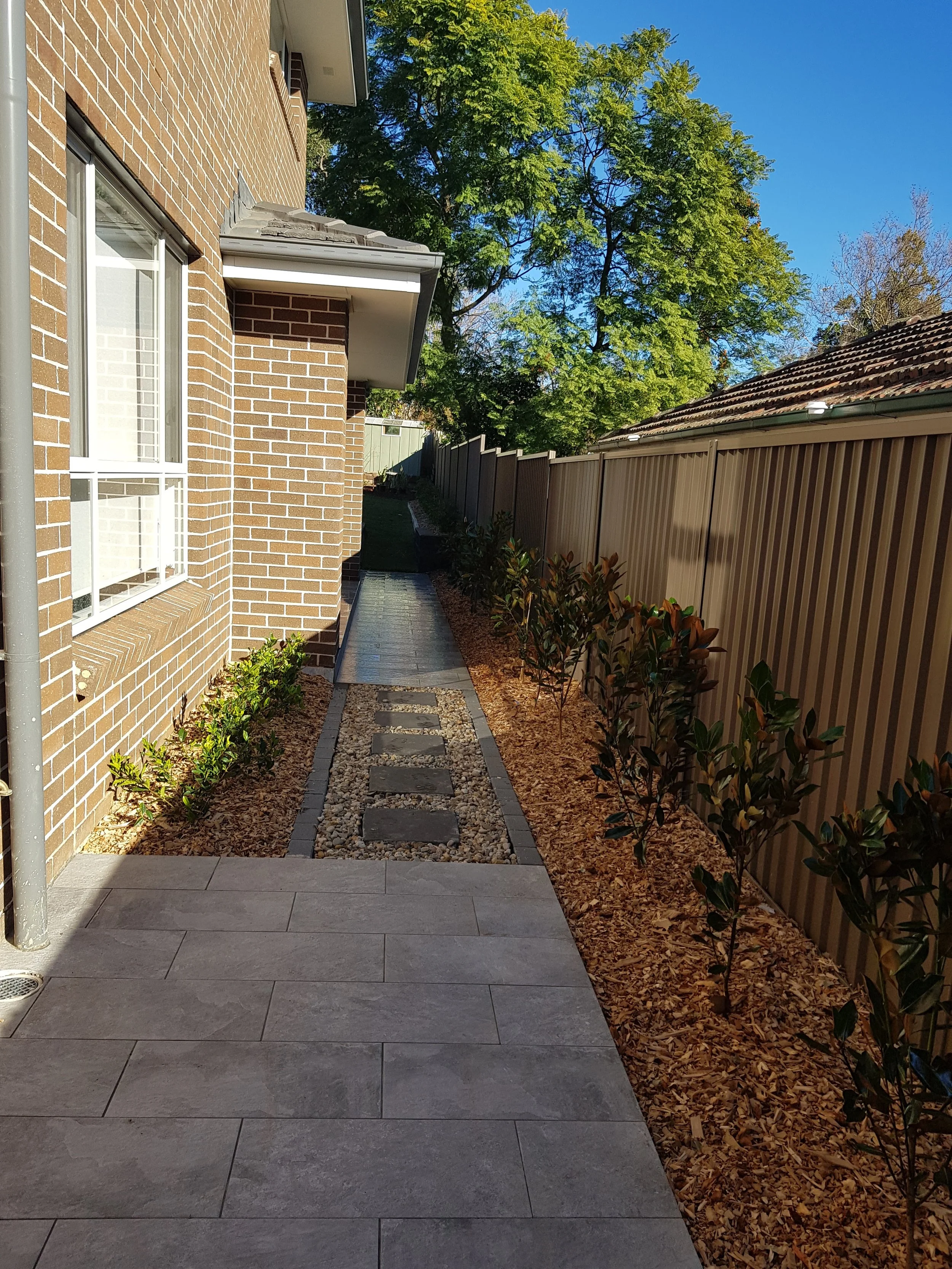 Side yard with a paved patio, small plants along the brick house, a pathway with stepping stones and pebbles, and a wooden fence enclosing the yard, under a bright blue sky.