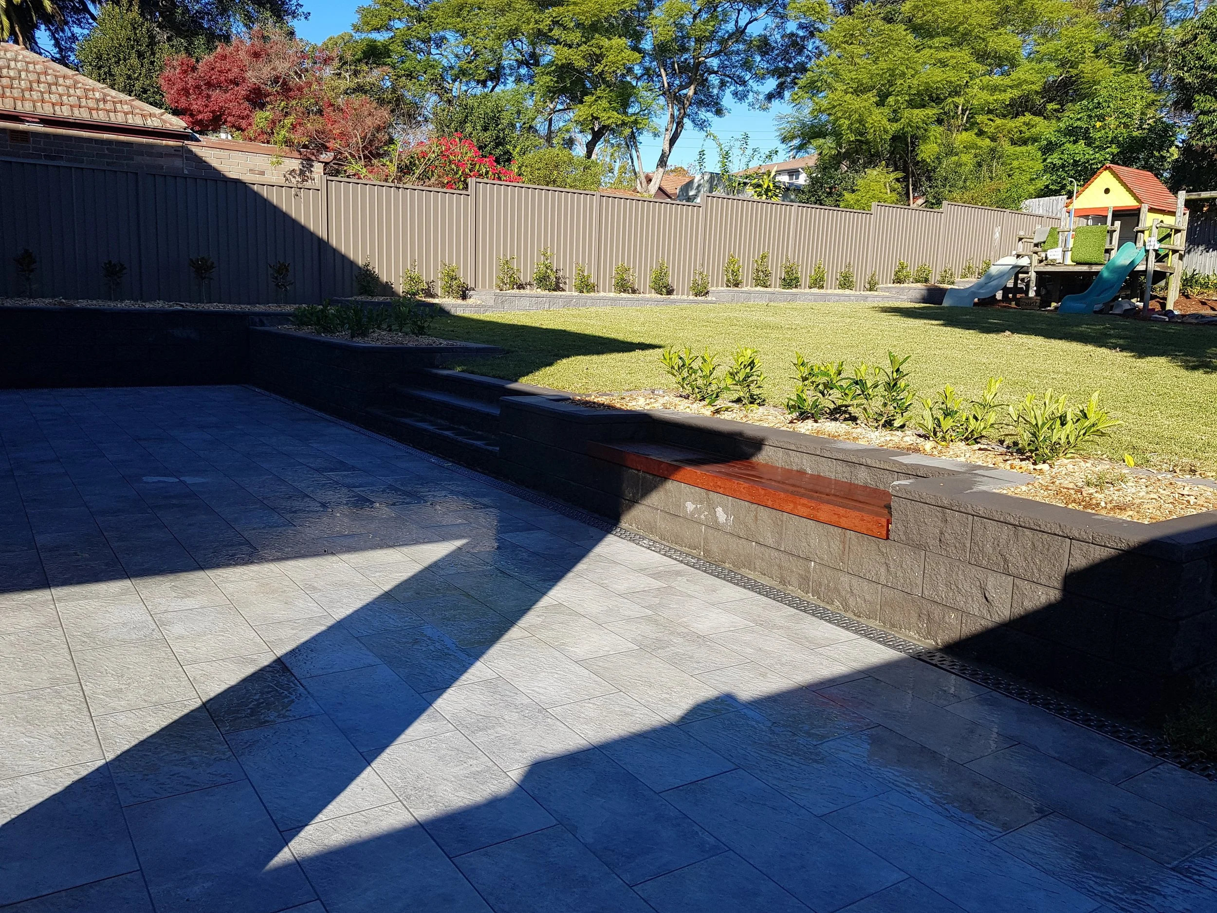 Backyard garden with a paved patio, a grassy lawn, a children’s playhouse with slides, and bushes along a fence, with trees in the background.