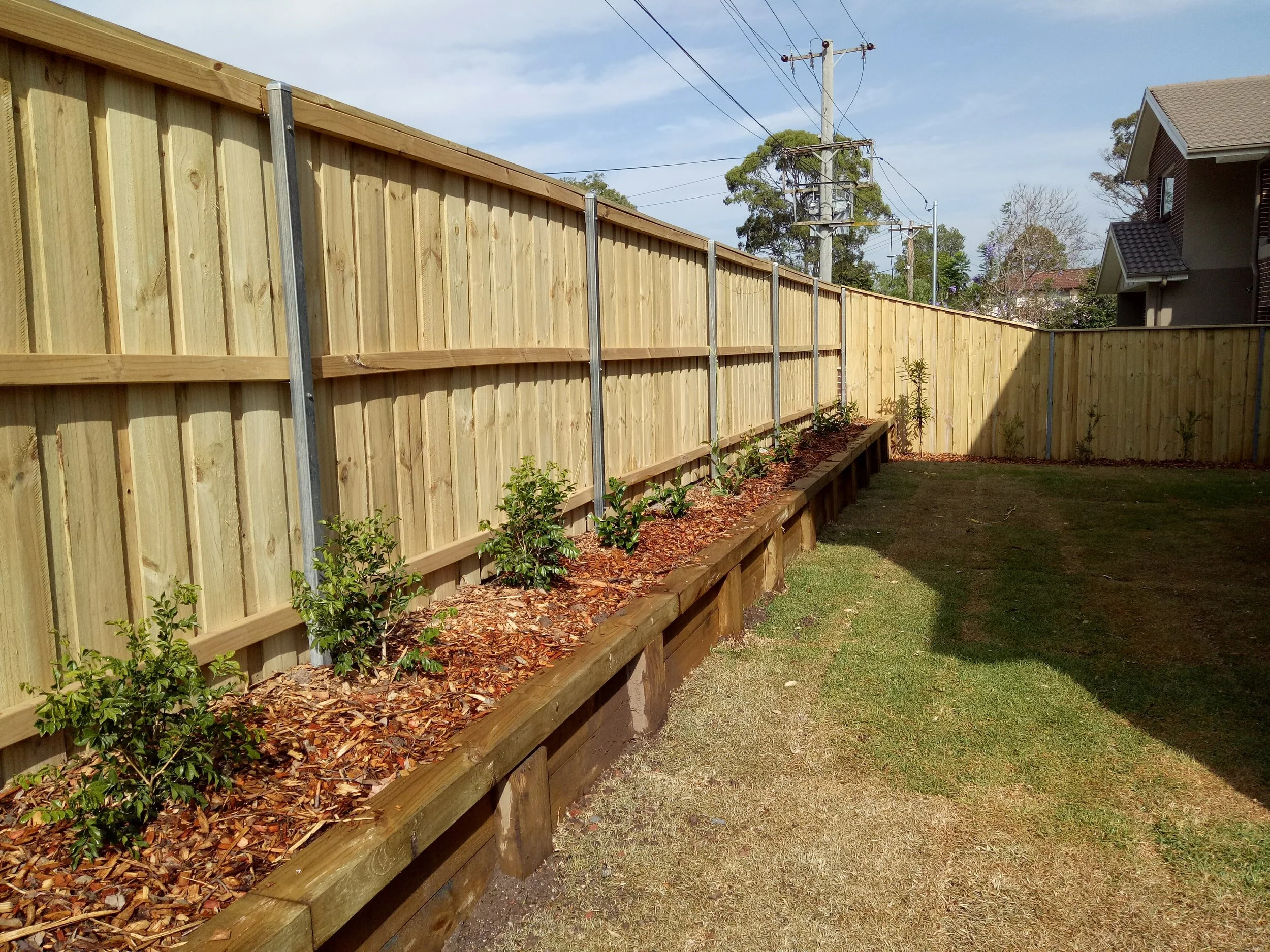 A backyard with a newly built wooden fence, a garden bed with small plants, green grass, and neighboring houses in the background under a partly cloudy sky.
