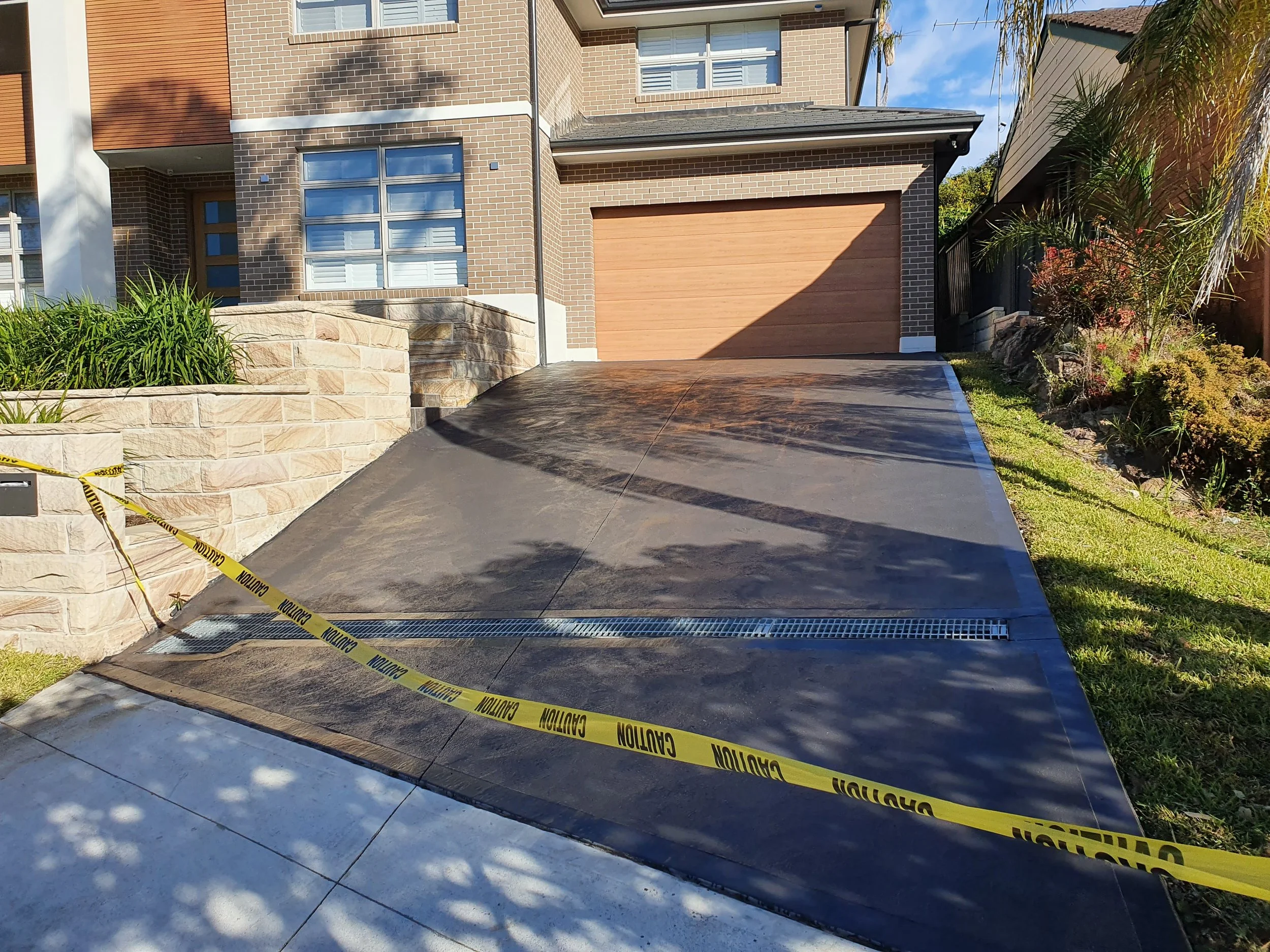 A newly sealed black asphalt driveway leading to a house with a brown garage door. The driveway is sectioned off with yellow caution tape. Surrounding the driveway are brick and stone walls, a grassy lawn, and landscaped plants.