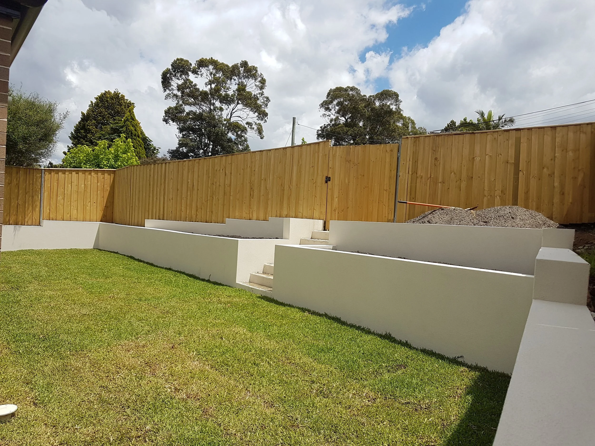 Newly built backyard with a vibrant green lawn, white retaining walls with steps, a wooden privacy fence, and partly cloudy sky.