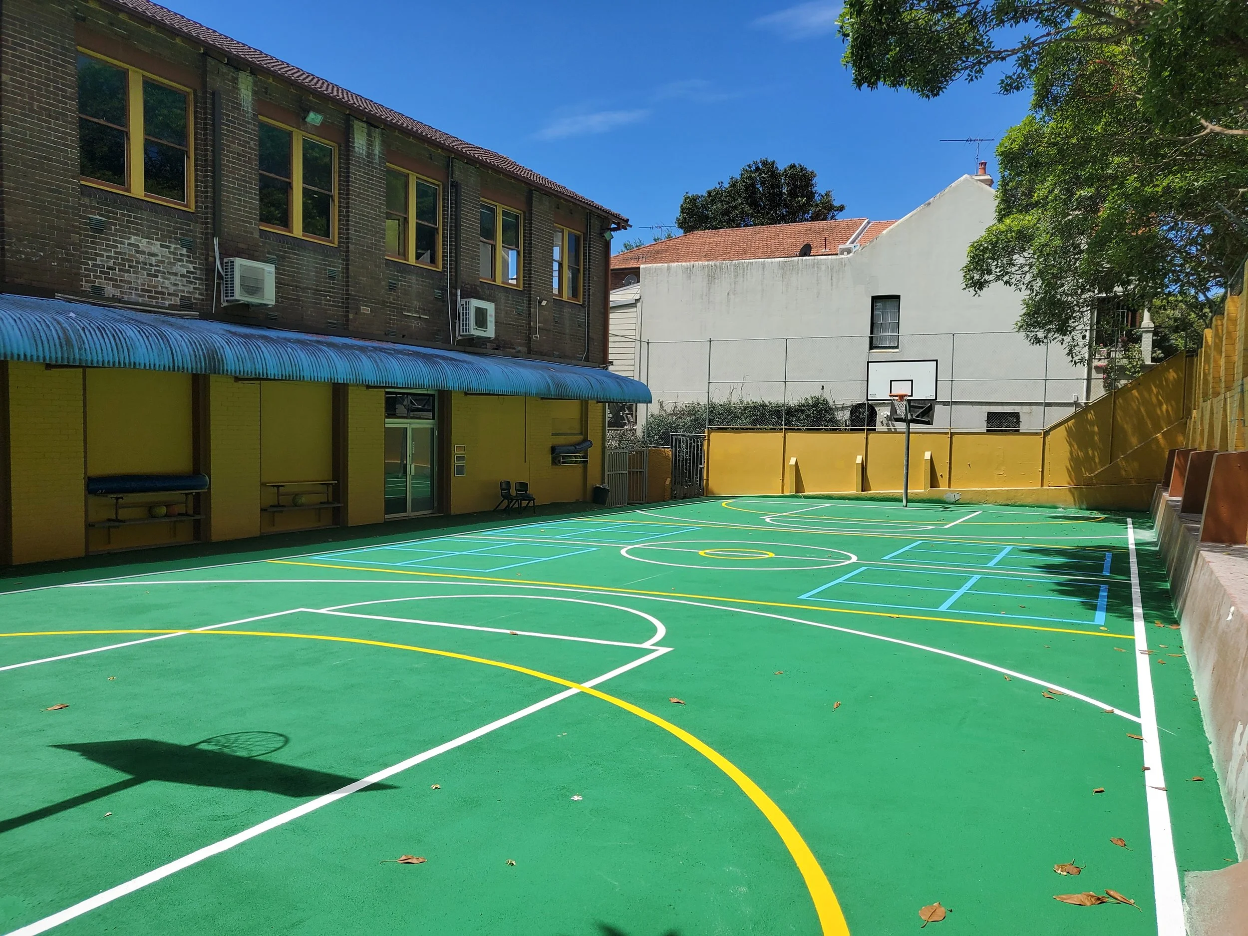 A colorful outdoor basketball court with yellow, white, and blue lines, surrounded by a yellow wall, with a basketball hoop on a pole and a bench, in a residential area with buildings and trees, under a clear blue sky.
