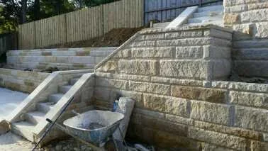Construction site with stone retaining wall and stairs, garden tools, and a wheelbarrow.