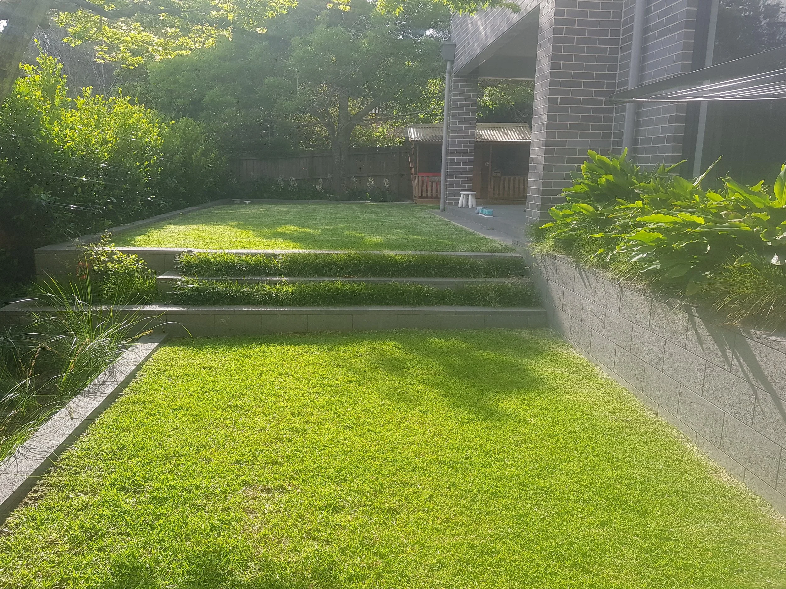 A neatly maintained backyard with a green lawn, a set of concrete steps leading to an elevated patio area, and lush plants along the edges. There's a brick house with an outdoor covered area, and a small shed in the background surrounding trees and a