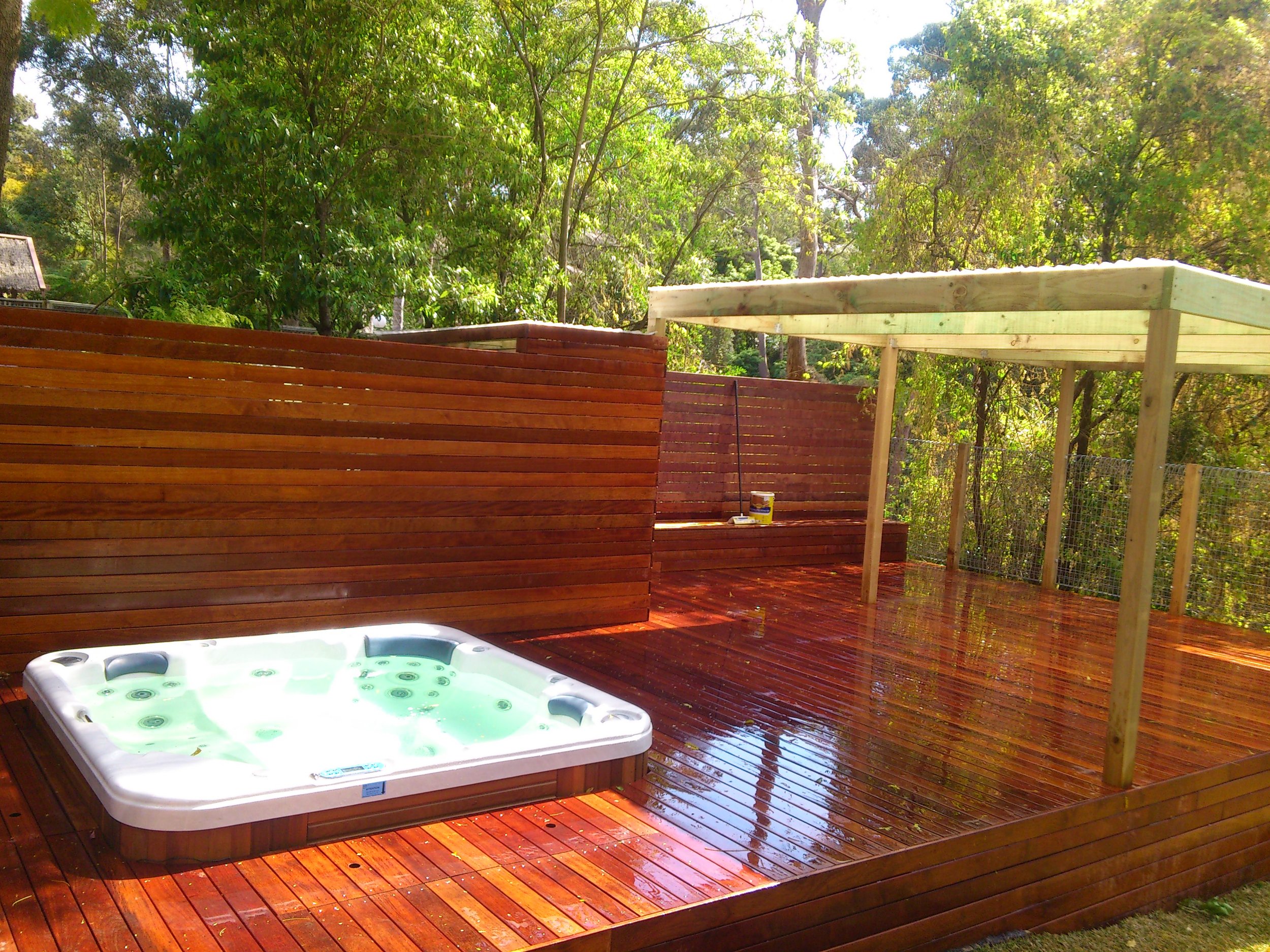 A wooden deck with a hot tub and a partially constructed shade structure in a backyard surrounded by trees.