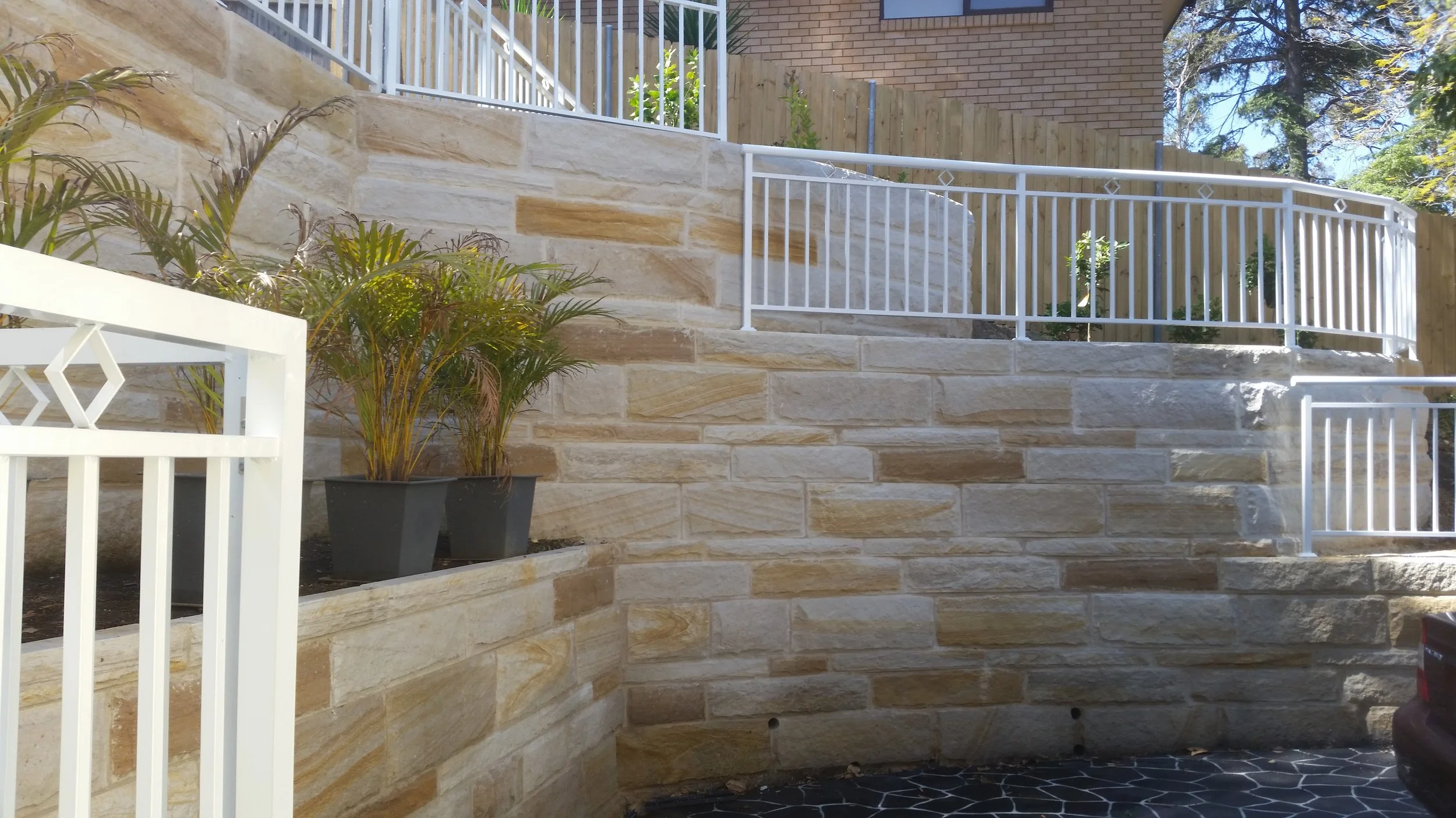 Retaining wall made of beige stone blocks with white metal railings on top, potted plants with green foliage, and a brick building in the background under a blue sky.