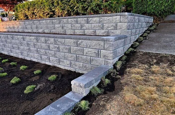 A stone retaining wall with neatly arranged bricks, bordered by small plants and soil, with a sidewalk casting shadows in the background.