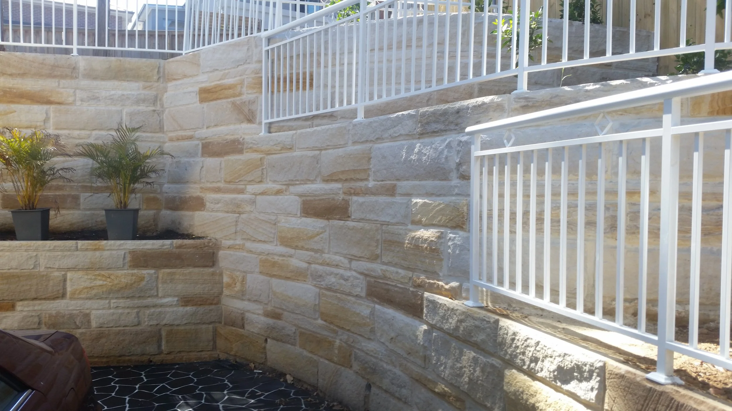 A tiered outdoor retaining wall made of beige stone blocks with white metal railings on each level. Two potted plants with green leaves are placed on the lower level, and the ground has black tiles with white patterns.