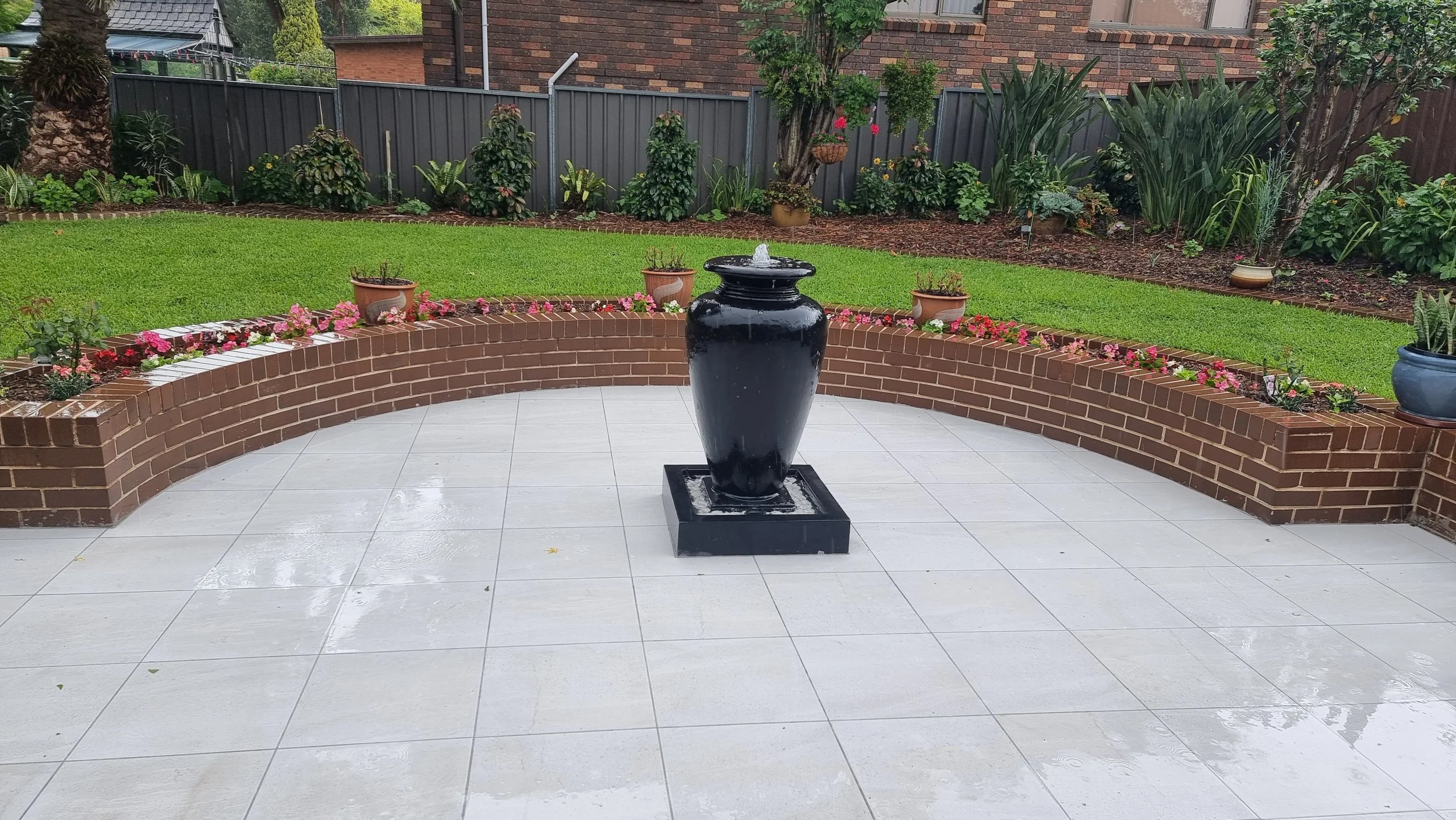 Black water fountain in the center of a patio, surrounded by a brick curved wall with flowers on top. Behind the wall is a garden with green grass, trees, and potted plants. The patio floor is made of light-colored tiles, wet from rain.