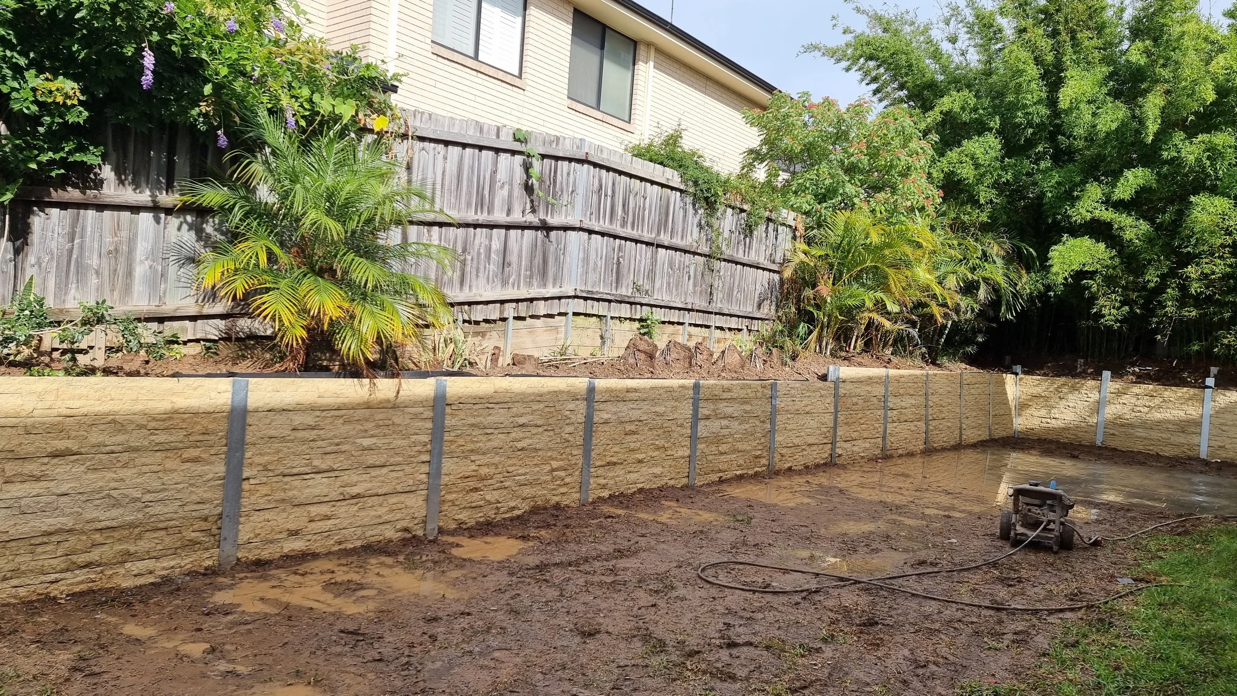 Backyard with a newly constructed retaining wall, muddy ground, a pressure washer, and green plants against a wooden fence and house.