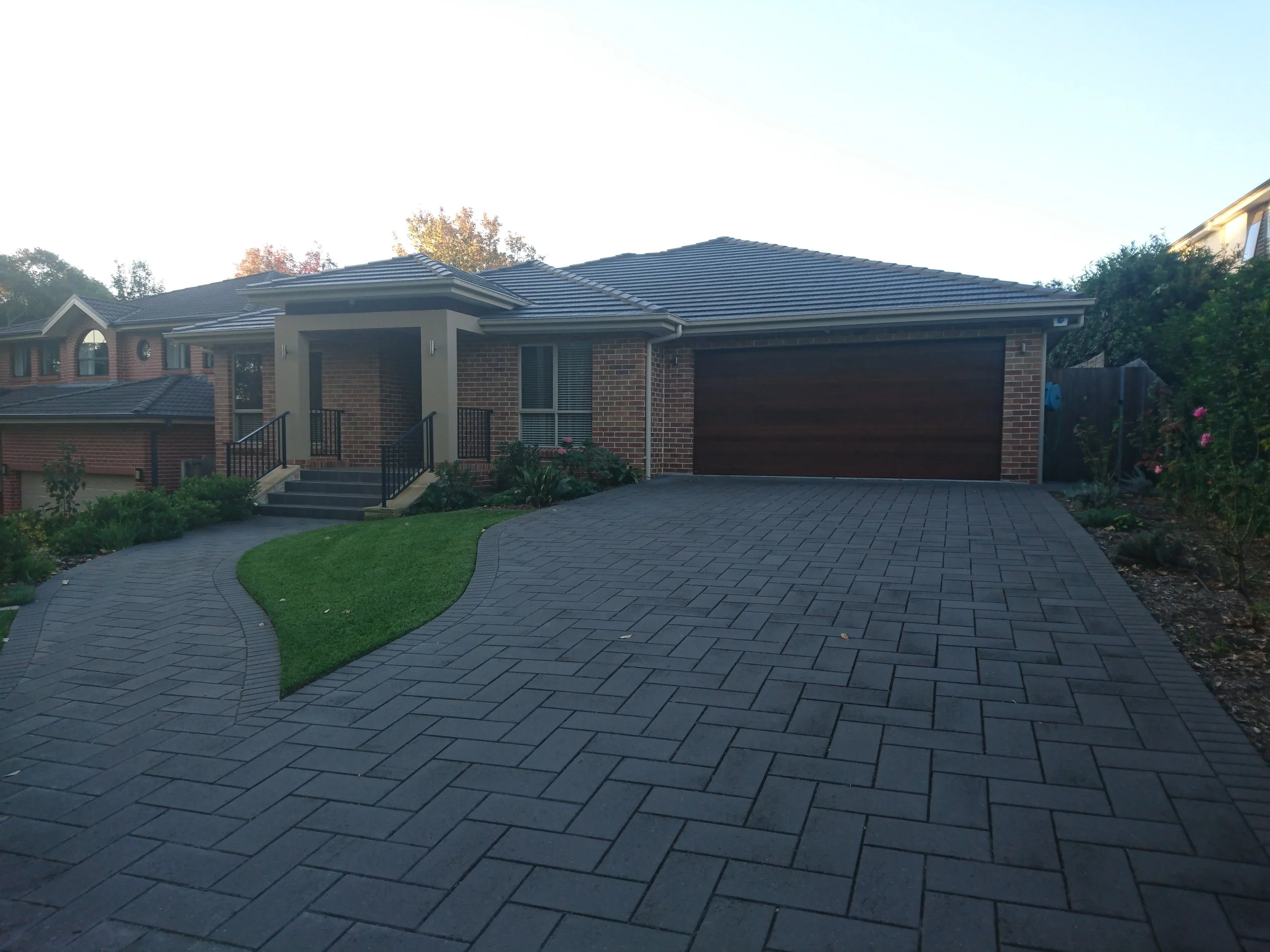 Modern brick house with a curved driveway, front steps, and a garage door. Well-maintained lawn and garden with flowering plants on the right side.
