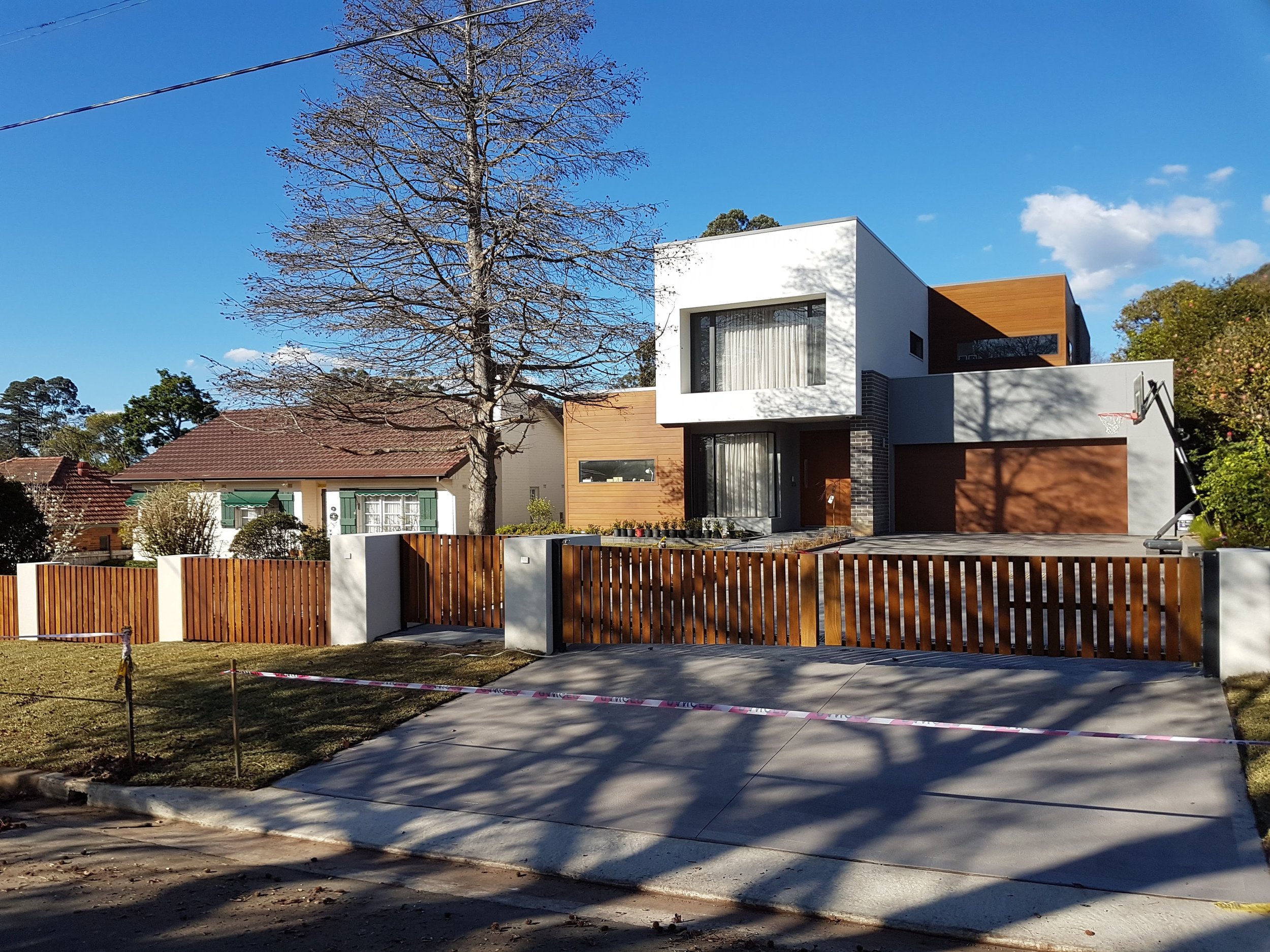 A modern two-story house with white, gray, and wood exterior, a large window, and a wooden fence in front. There is a leafless tree in the yard with a concrete driveway and a small basketball hoop attached to the house.