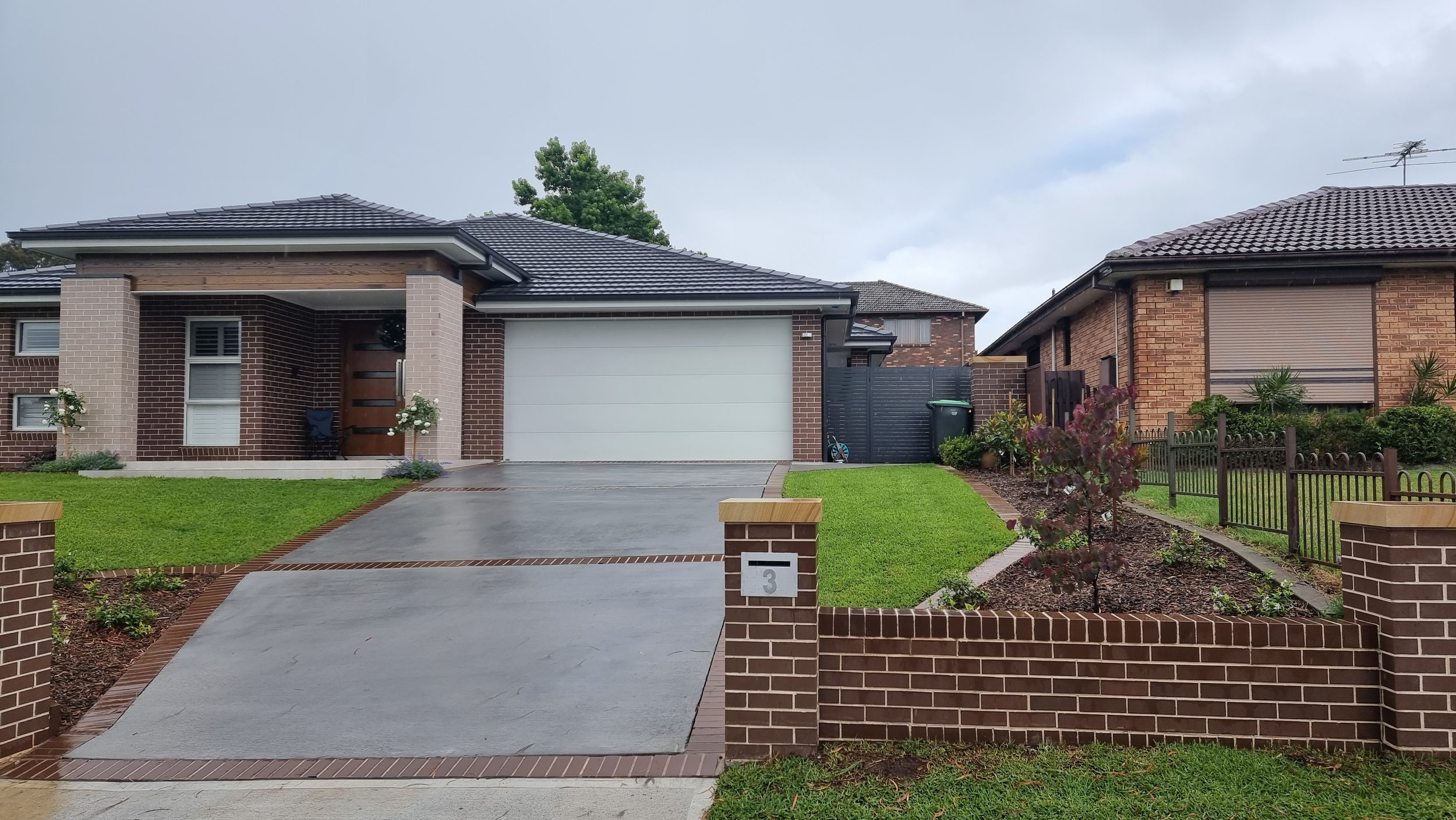 A modern brick house with a gray garage door, a concrete driveway, and a small front garden with green grass and a young tree. There is a brick fence with the house number 3 on a brick pillar.