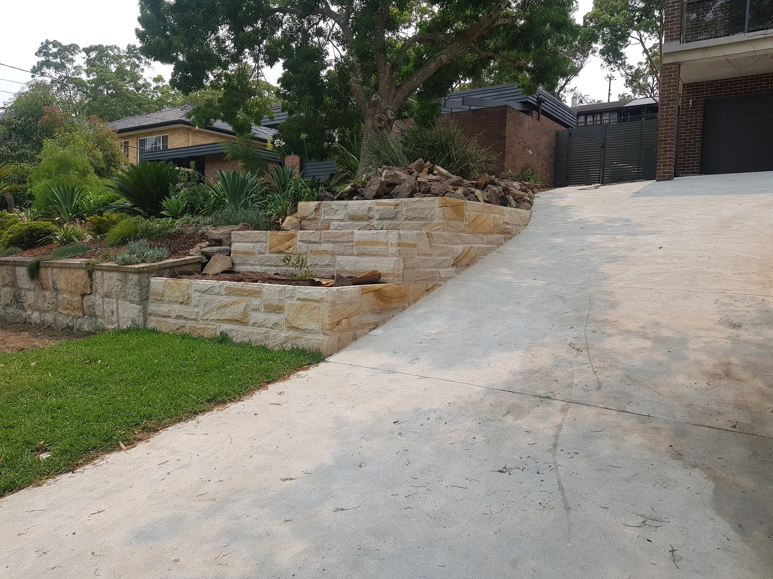 Concrete driveway sloping uphill next to a landscaped garden with various plants and a retaining wall made of beige stone bricks in front of a house with brick walls and dark window frames.