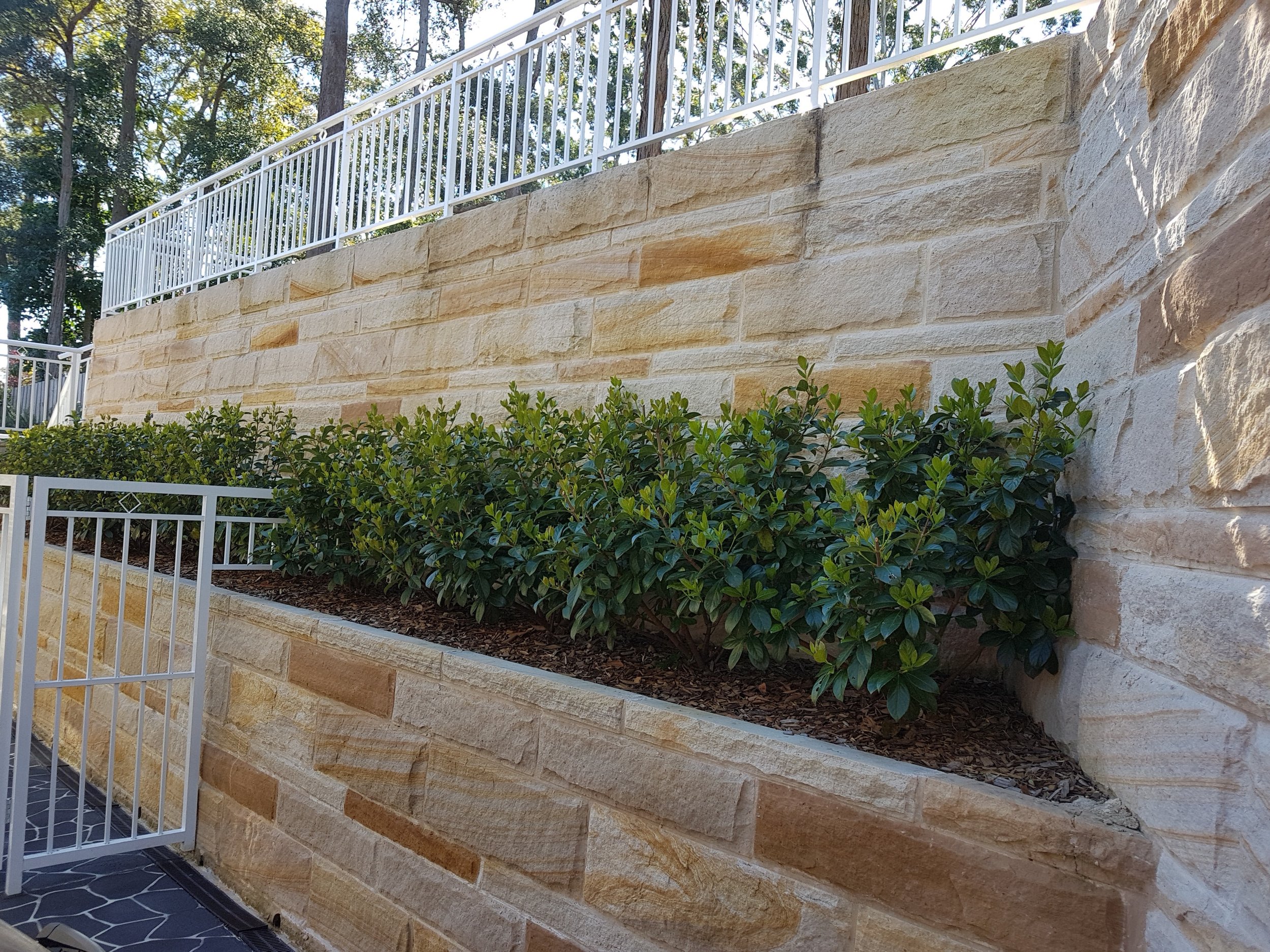 A landscaped outdoor area with a tiered stone retaining wall, green bushes, and white metal railings.