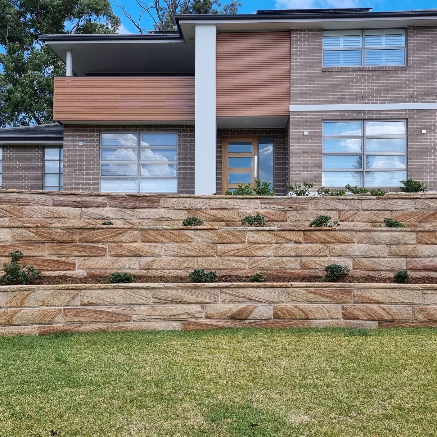 Modern multi-story house with brick facade, wooden and white accents, large windows reflecting blue sky and clouds, balcony on upper floor, stone retaining wall with small plants, green lawn in foreground.
