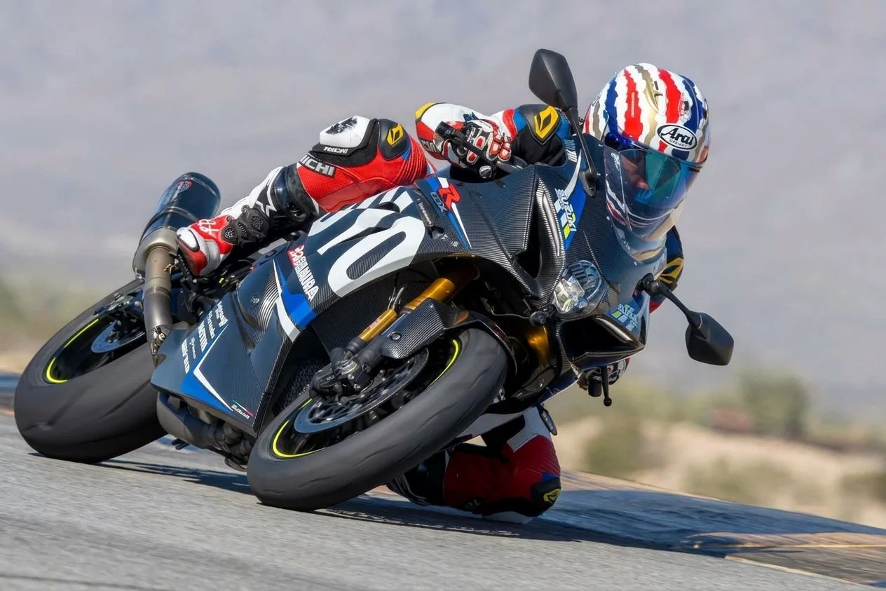 Motorcycle racer leaning into a turn on a race track, wearing a black, red, and yellow helmet and a racing suit with red, black, and gray colors, on a black and blue sport bike.