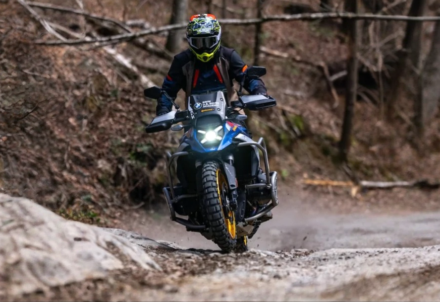 Motorcycle rider wearing a helmet and riding on a rugged dirt trail in a wooded area.