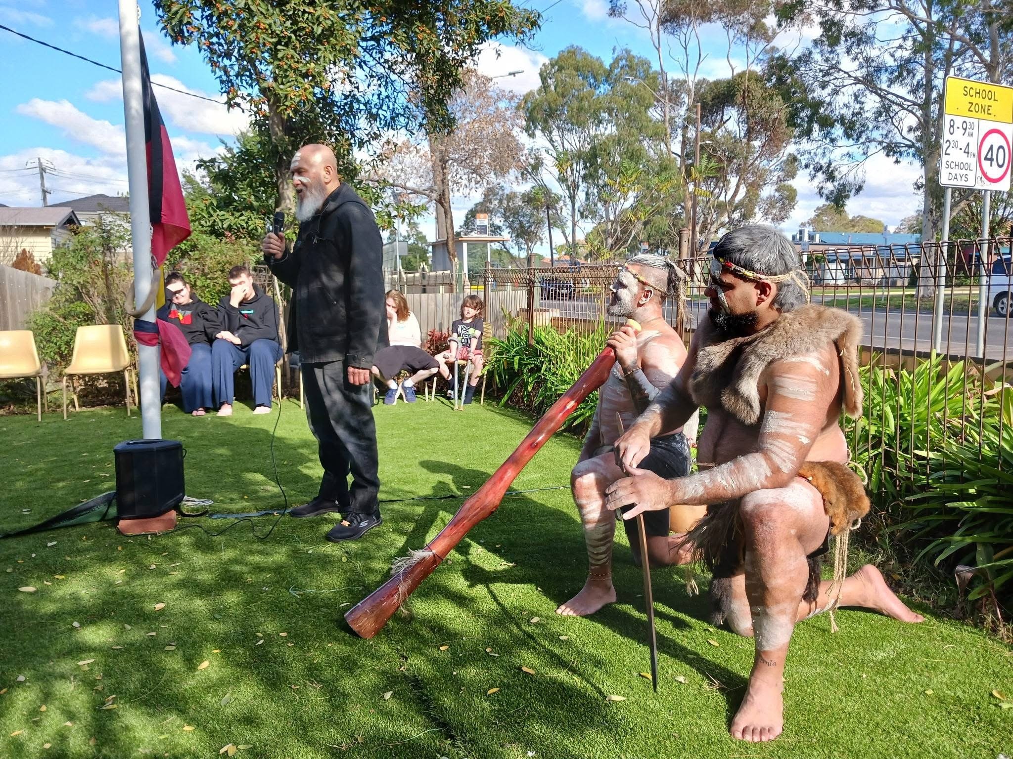 A group of people dressed as Indigenous Australians, sitting and standing in a park, with two standing men with painted faces, one holding a spear, and a man with a beard and whistle speaking into a microphone. There are children and other spectators