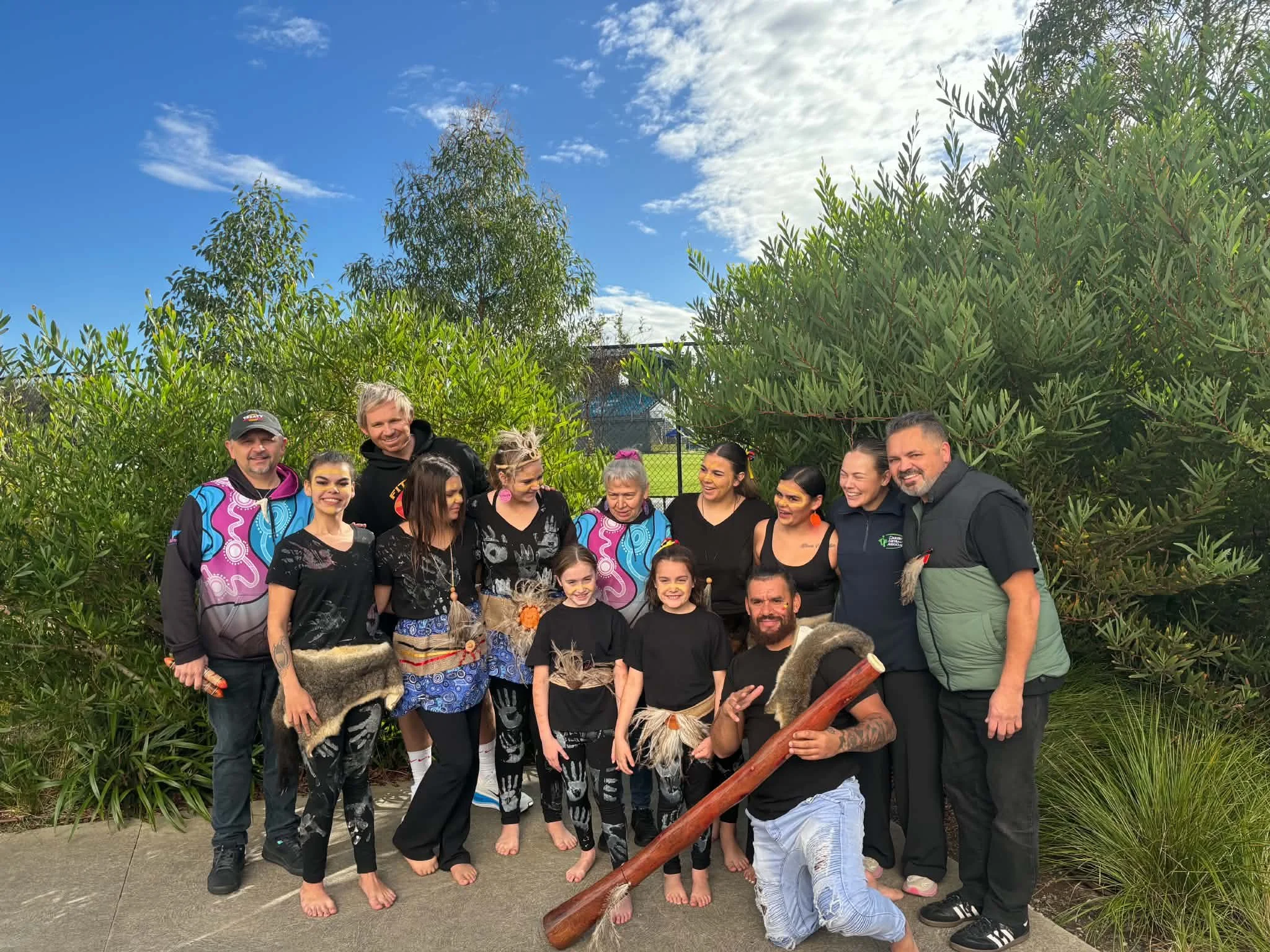 Group of people outdoors, some dressed in traditional indigenous clothing, surrounded by greenery with trees and a blue sky with some clouds.