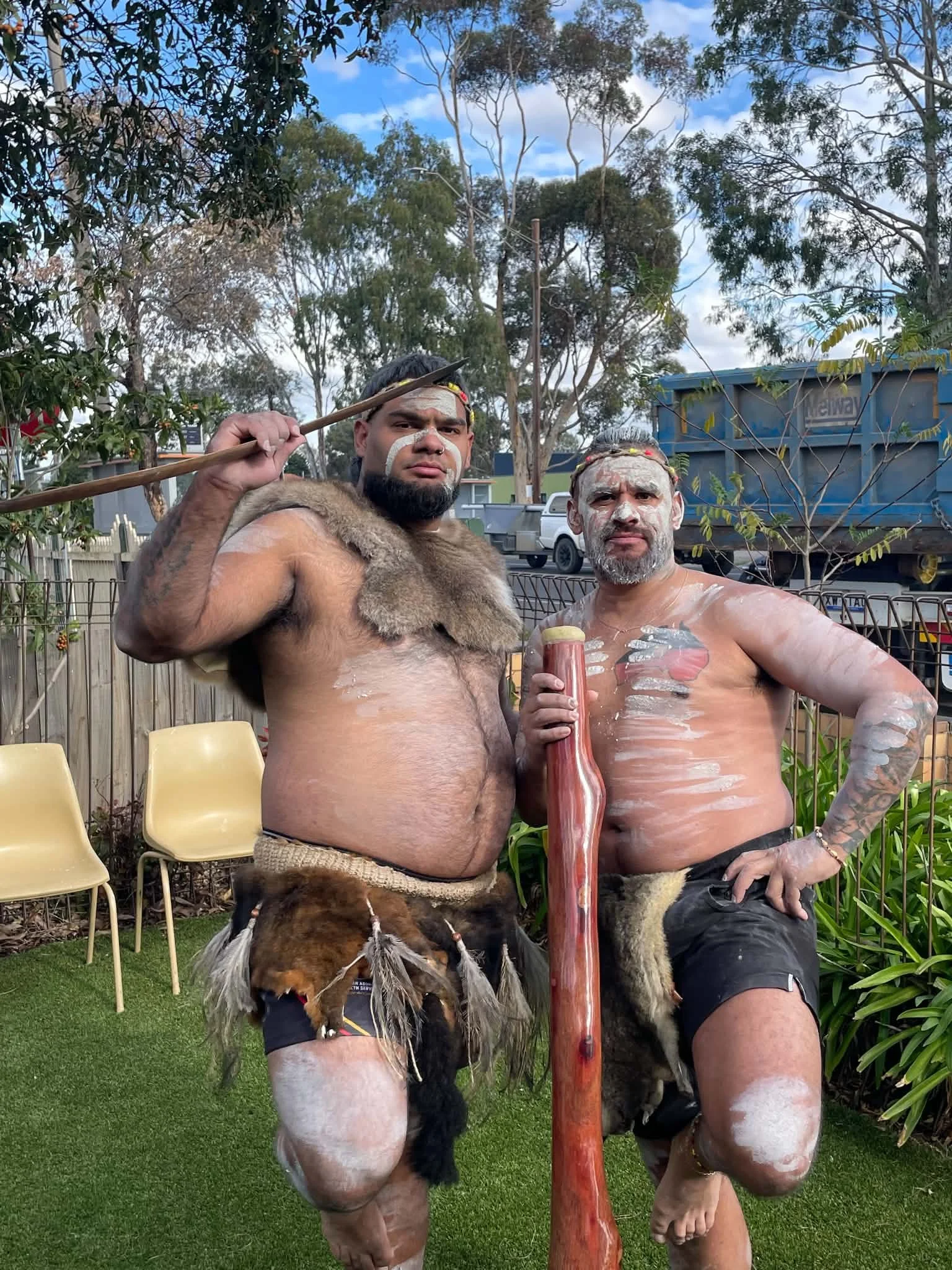 Two men with traditional face paint and body paint, wearing only shorts, holding a large carved wooden object, surrounded by dogs and outdoor furniture, in a backyard setting with trees and a blue sky.