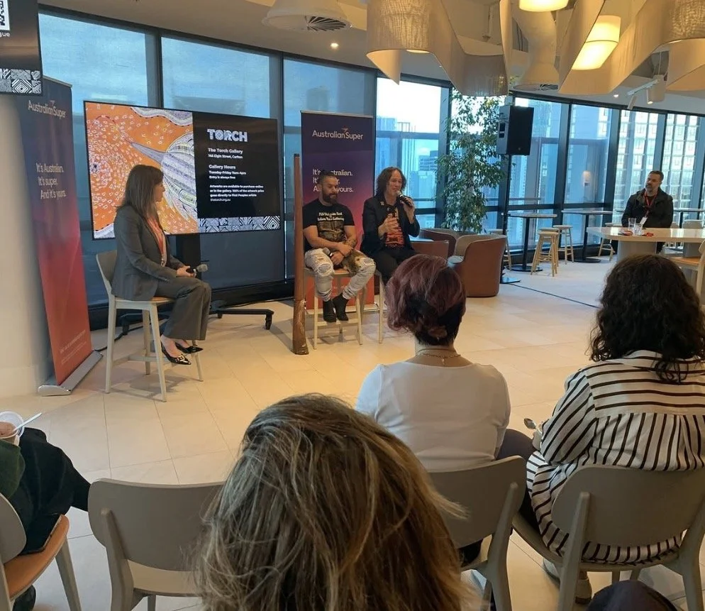A panel discussion taking place in a modern indoor venue with floor-to-ceiling windows. Four people are seated on stage, with three engaging in a conversation and one person listening. An audience is watching, seated on chairs. A large screen displays information about the event, and banners for AustLian Super and The Torch Gallery are visible.