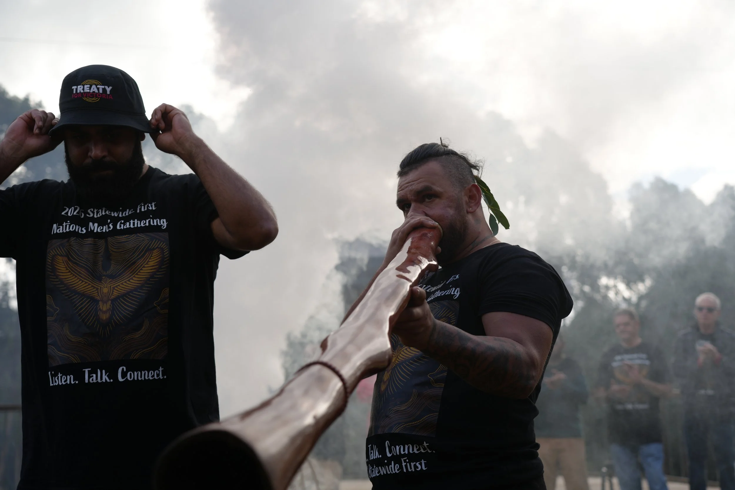 Two men participating in a culturally significant event with smoke and trees in the background. One man is adjusting a black hat, and the other is drinking from a long wooden pipe. Both are wearing black shirts with text related to a nation’s gathering.