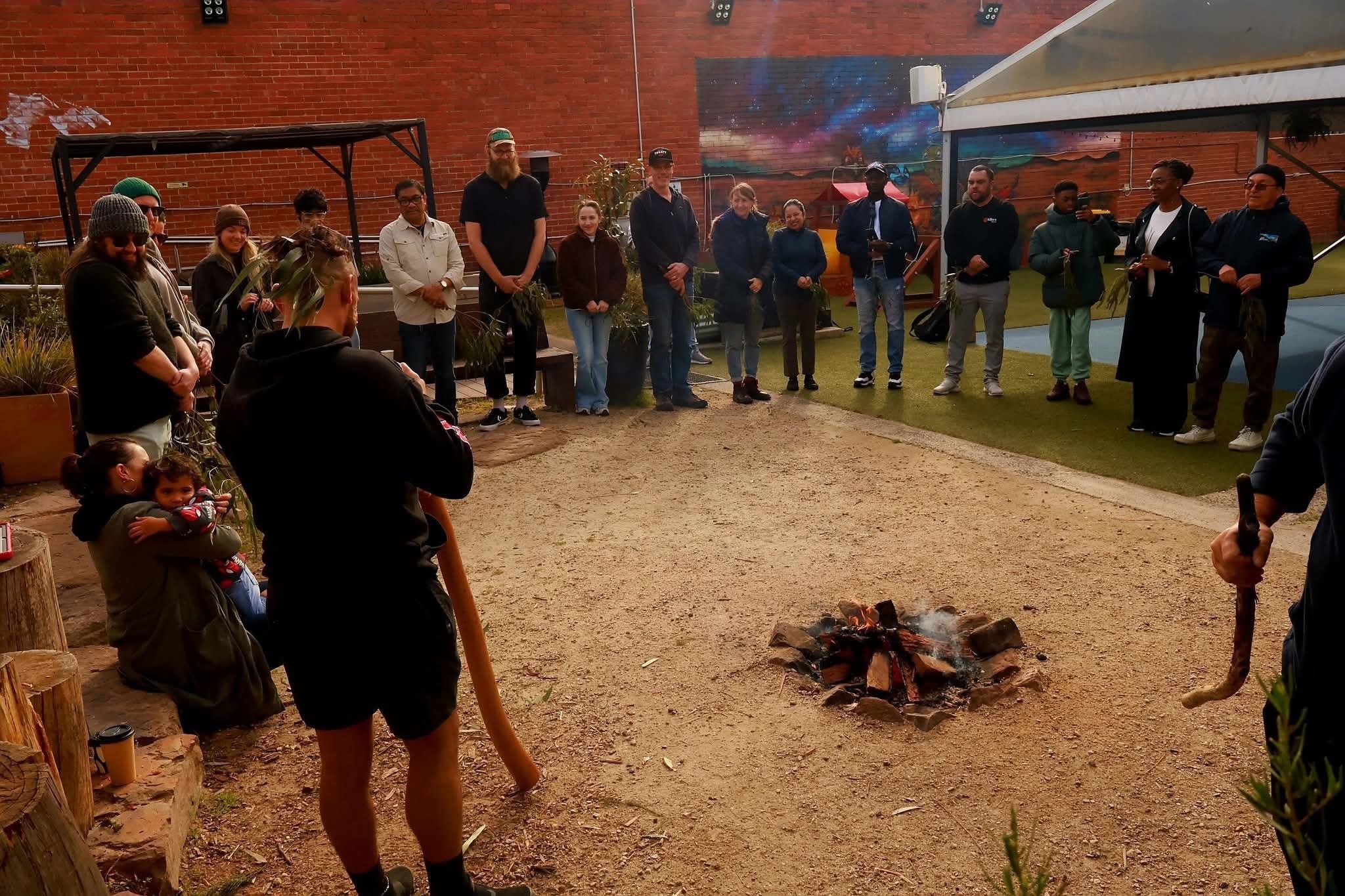A group of people gathered outdoors around a small campfire on a Dirt area, with some holding plants and others taking photos, in front of a brick wall and colorful mural.